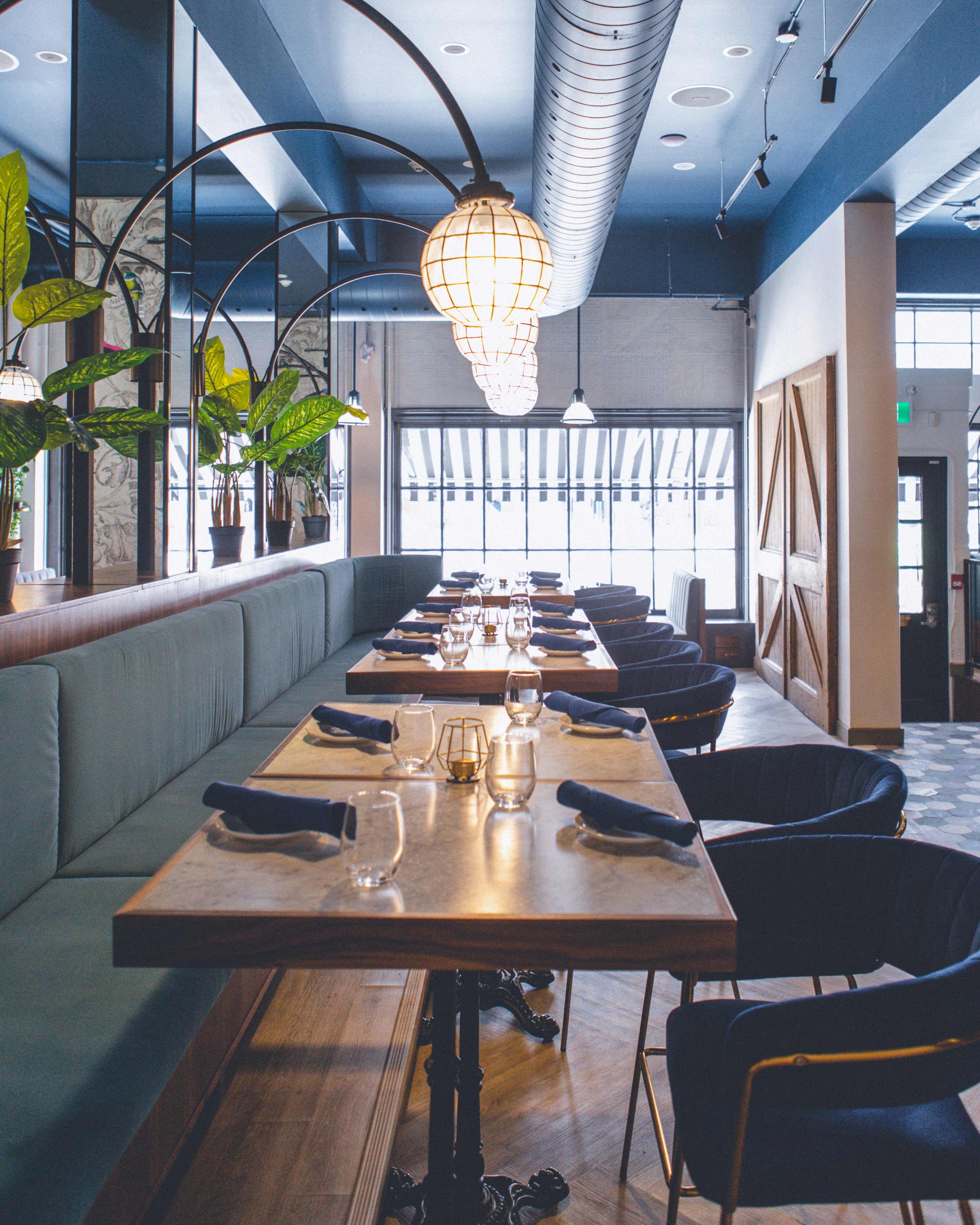 A modern restaurant interior with a long wooden table set for dining, surrounded by black chairs. The table is adorned with glasses, napkins, and small decorative items. There is a green cushioned bench along one side of the table and a row of potted