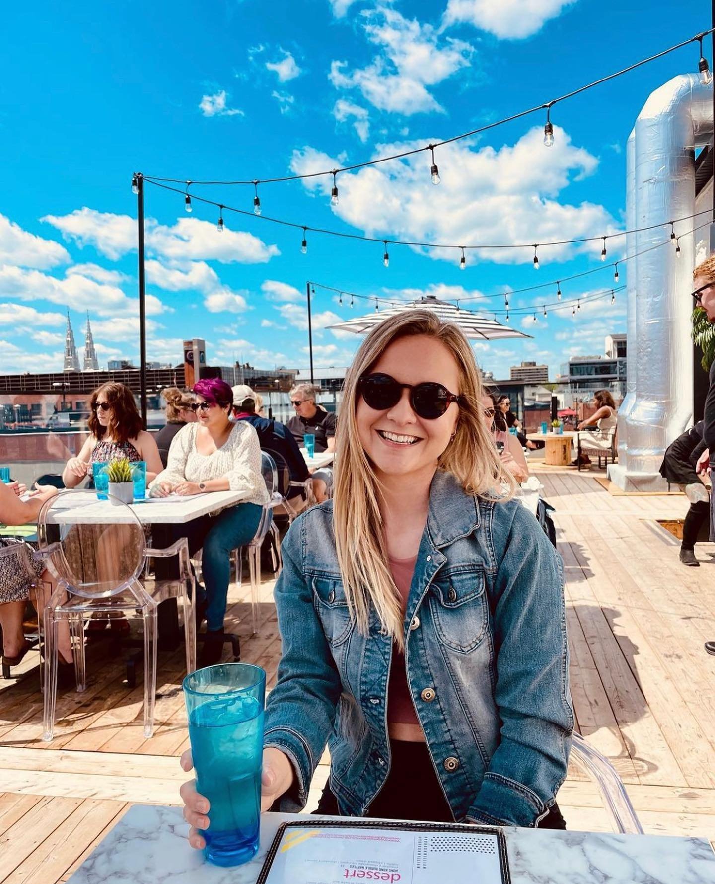 A young woman with blonde hair and sunglasses smiling and holding a blue drink at an outdoor rooftop restaurant on a sunny day.