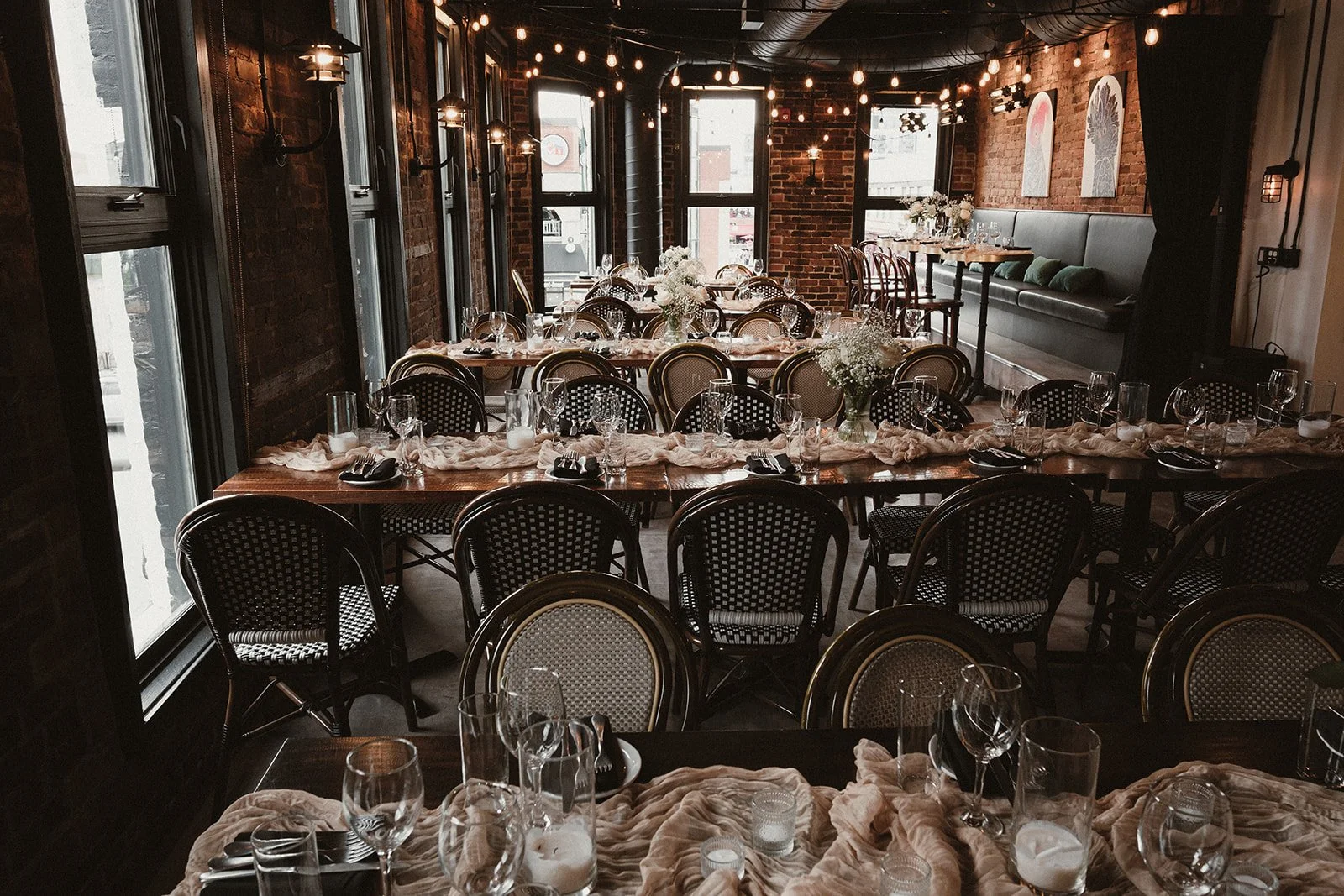 Elegant dining setup with long wooden tables decorated with beige table runners, glass vases filled with white flowers, and set with glassware and black napkins. The room has exposed brick walls, large windows, and string lights hanging from the ceil
