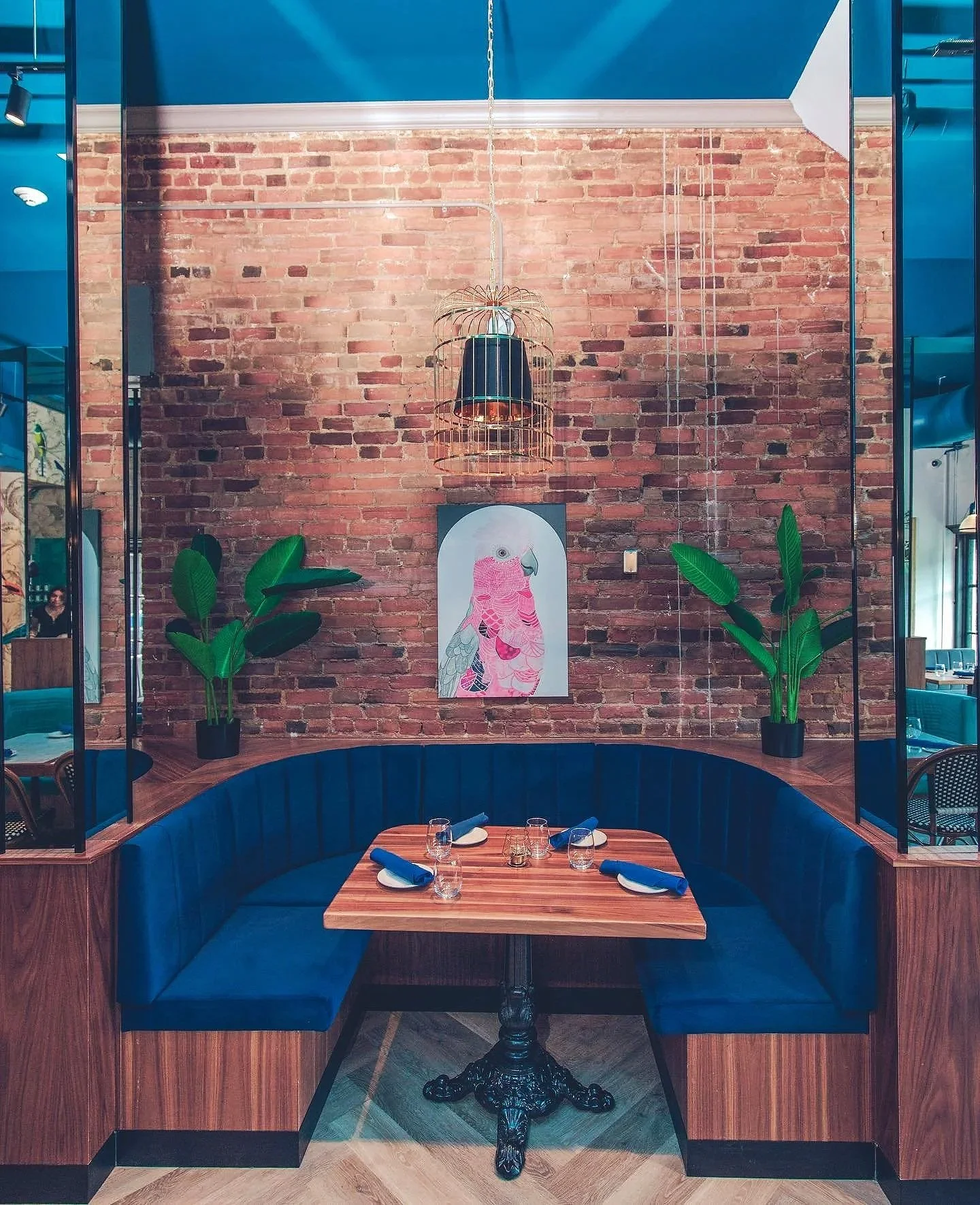 Restaurant velvet booth with a wooden table set with glasses and napkins, blue cushioned seating, two large green potted plants, brick wall with a pink flamingo artwork, hanging cage-style light fixture, and blue ceiling. modern venue events parties