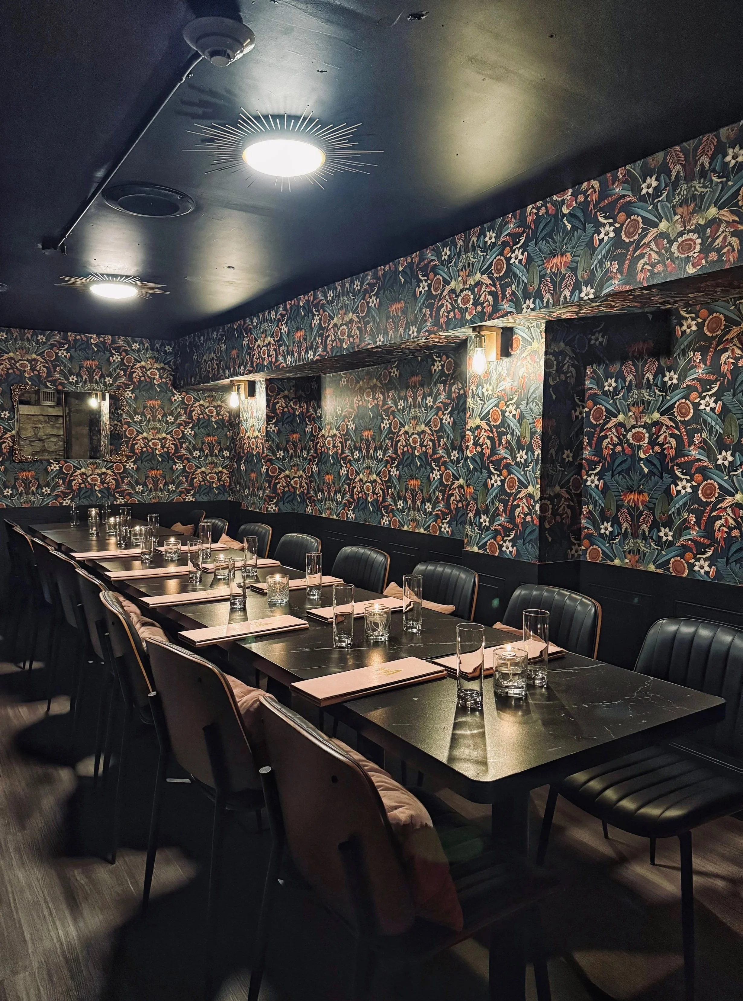 A long rectangular dining table set with glasses, menus, and napkins in a dimly lit restaurant with dark chairs, floral wallpaper, and ceiling lights. Best bar in Ottawa. Speakeasy room behind the bar. 