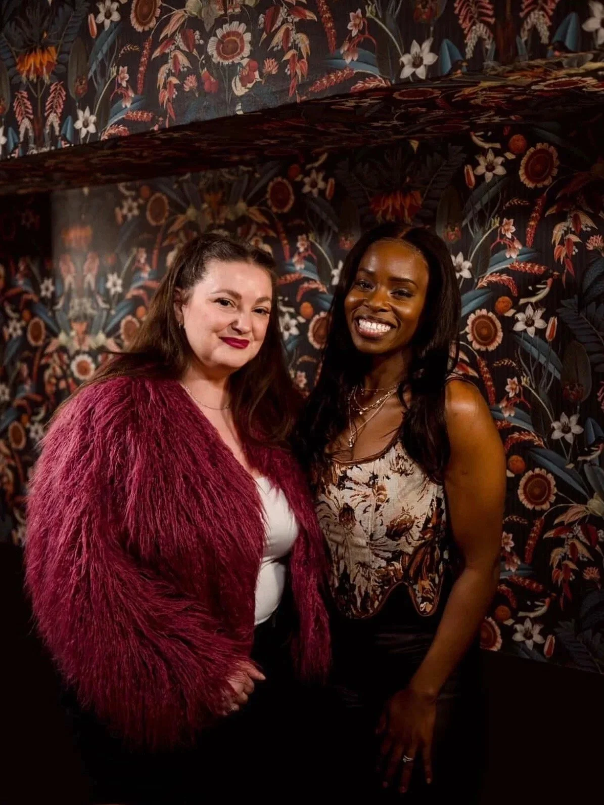Two women standing in front of a dark floral-patterned wallpaper. The woman on the left has long brown hair, wears a white top and a red fuzzy jacket. The woman on the right has long black hair, wears a patterned sleeveless top and is smiling.