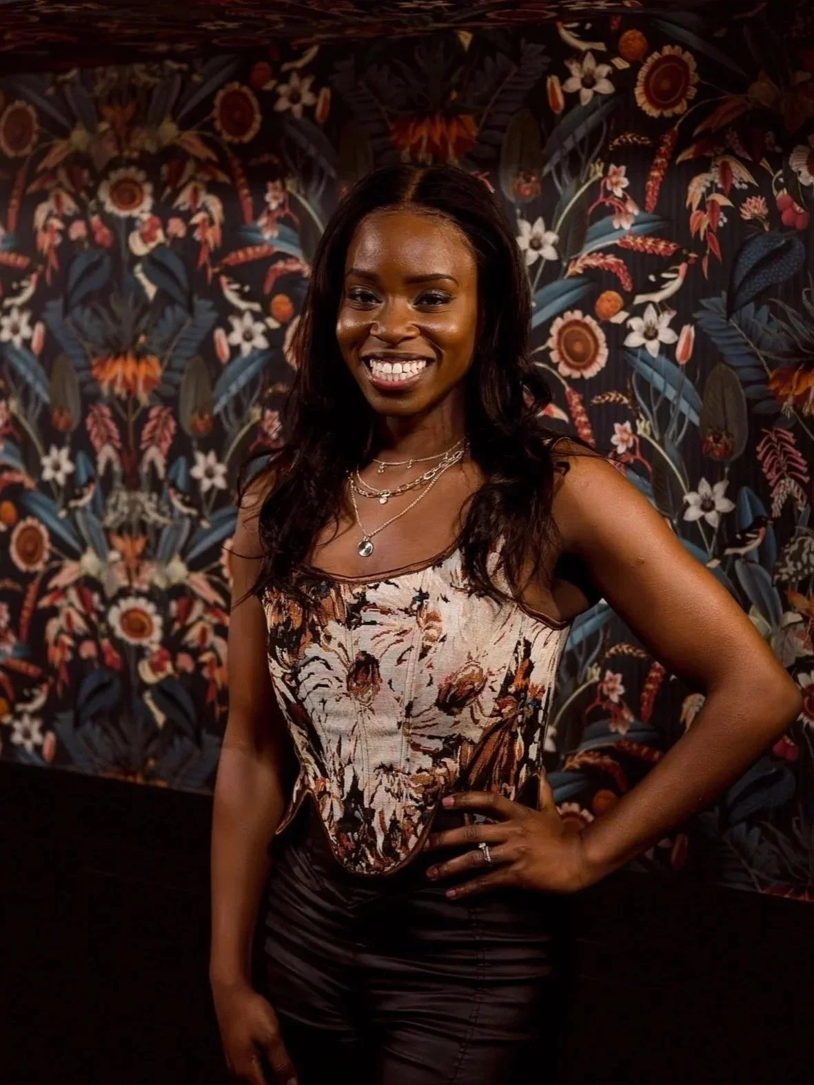 A woman with long dark wavy hair smiling and posing with her hand on her hip in front of a colorful, floral-patterned wall. speakeasy private room Ottawa venues 18 people
