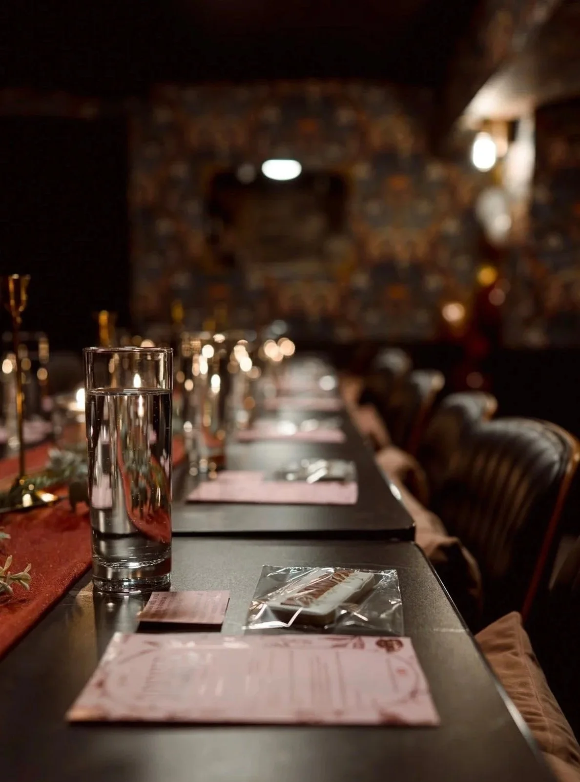 Table set with pink menus, a glass of water, and utensils in a dimly lit restaurant with leather chairs and a patterned wall background. Elegant setting, velvet curtains, floral wallpaper, heritage space, private bookings, corporate events 