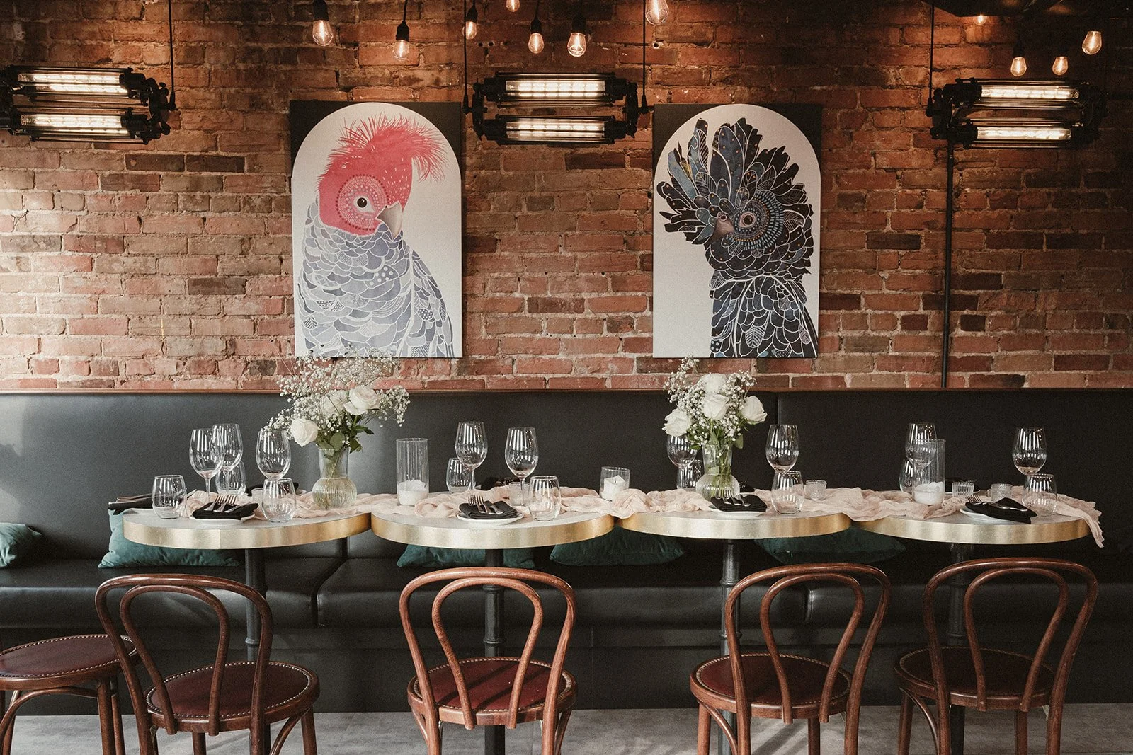 Restaurant table set for dining with wine glasses, floral centerpieces, and black napkins. Artistic animal portraits hang on a brick wall behind. Wooden chairs surround the table.