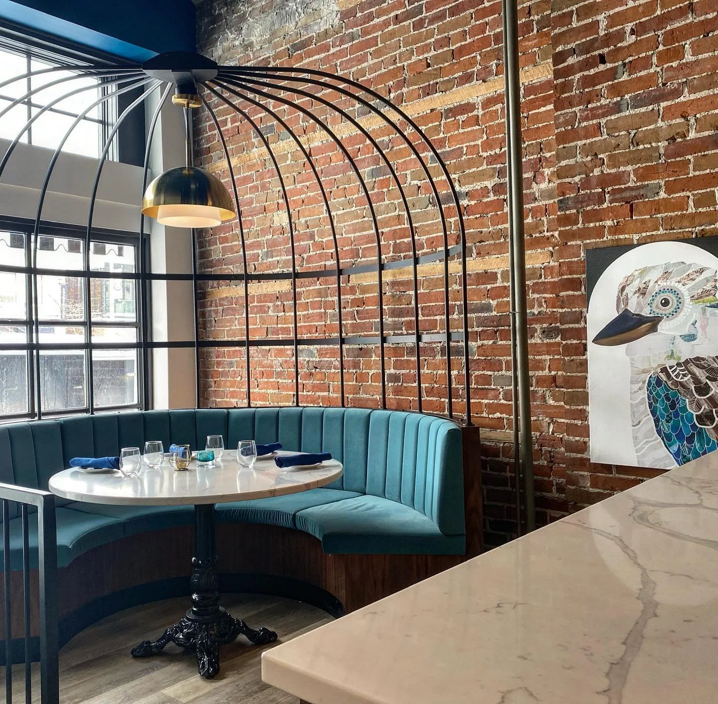 A corner of a restaurant with a round table set for four, a teal curved banquette, exposed brick wall, large window, and decorative lighting, with a colorful artwork of a bird on the wall.