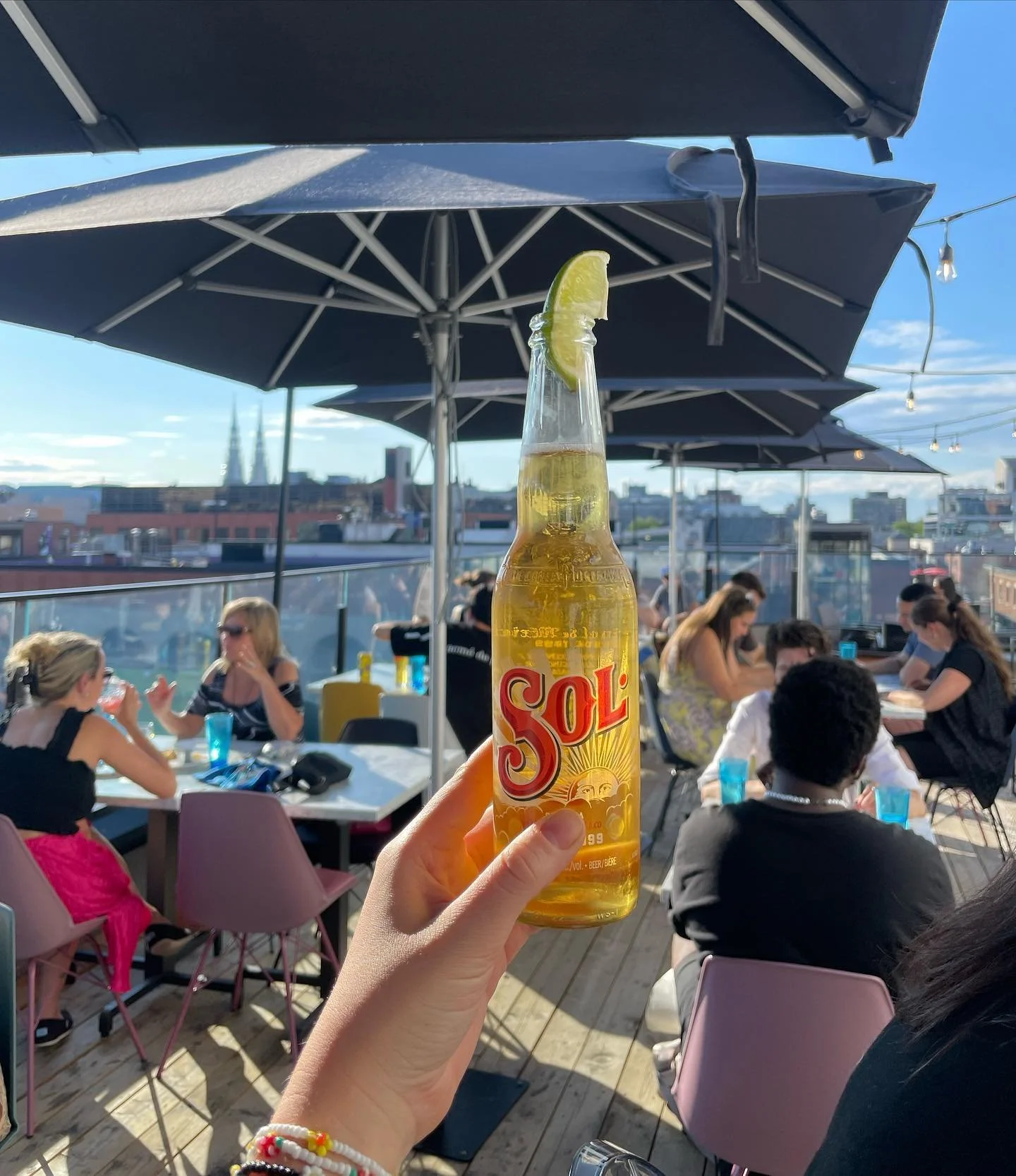 A person holding a Sol beer with a lime wedge on a rooftop patio during daytime, with groups of people seated at tables under umbrellas and a city skyline in the background.