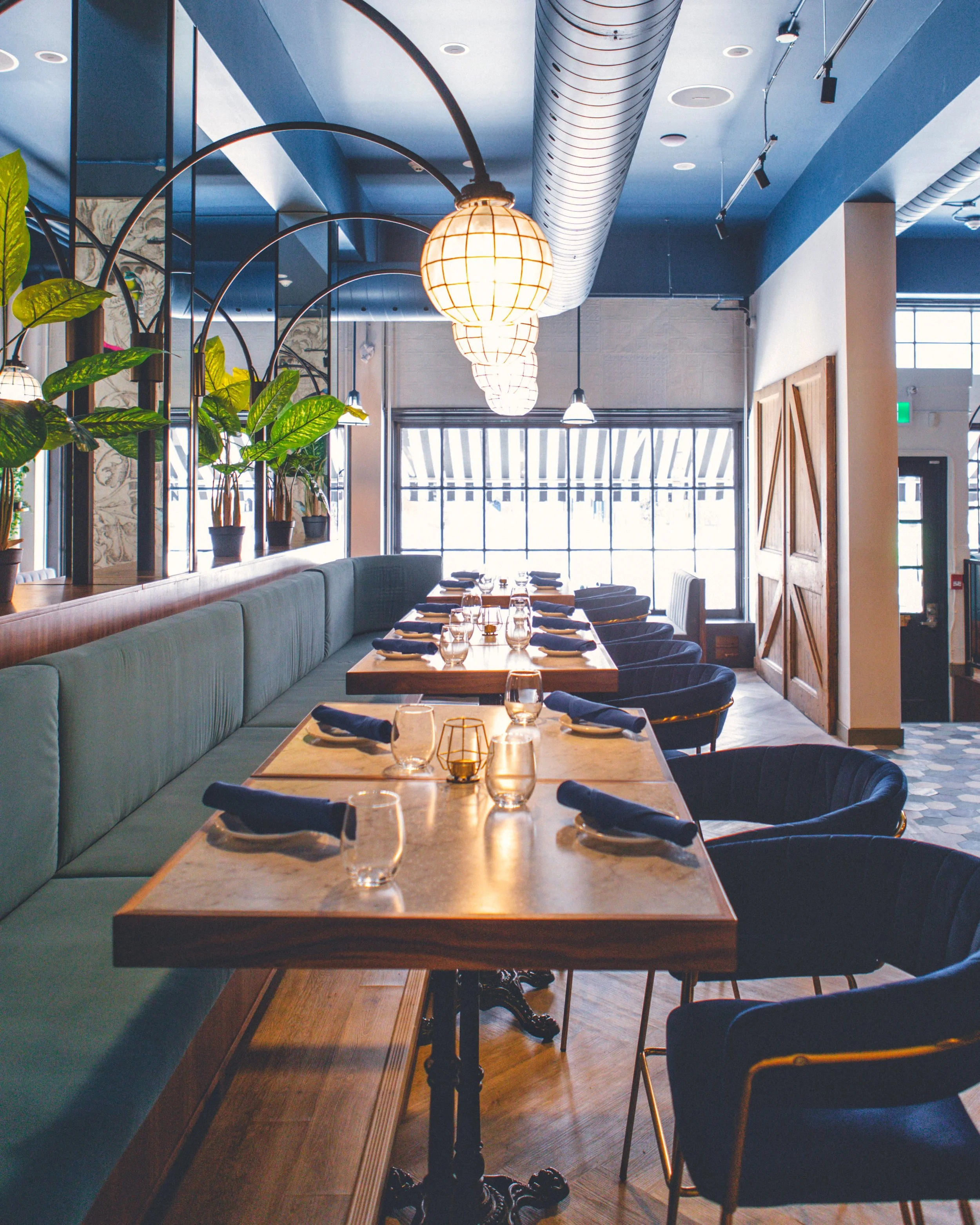 Interior of a restaurant with a long wooden table set with glasses, plates, and utensils. There is a green cushioned bench along one side and black chairs on the opposite side. Pendant lights hang from the ceiling, and potted plants are on a shelf. L