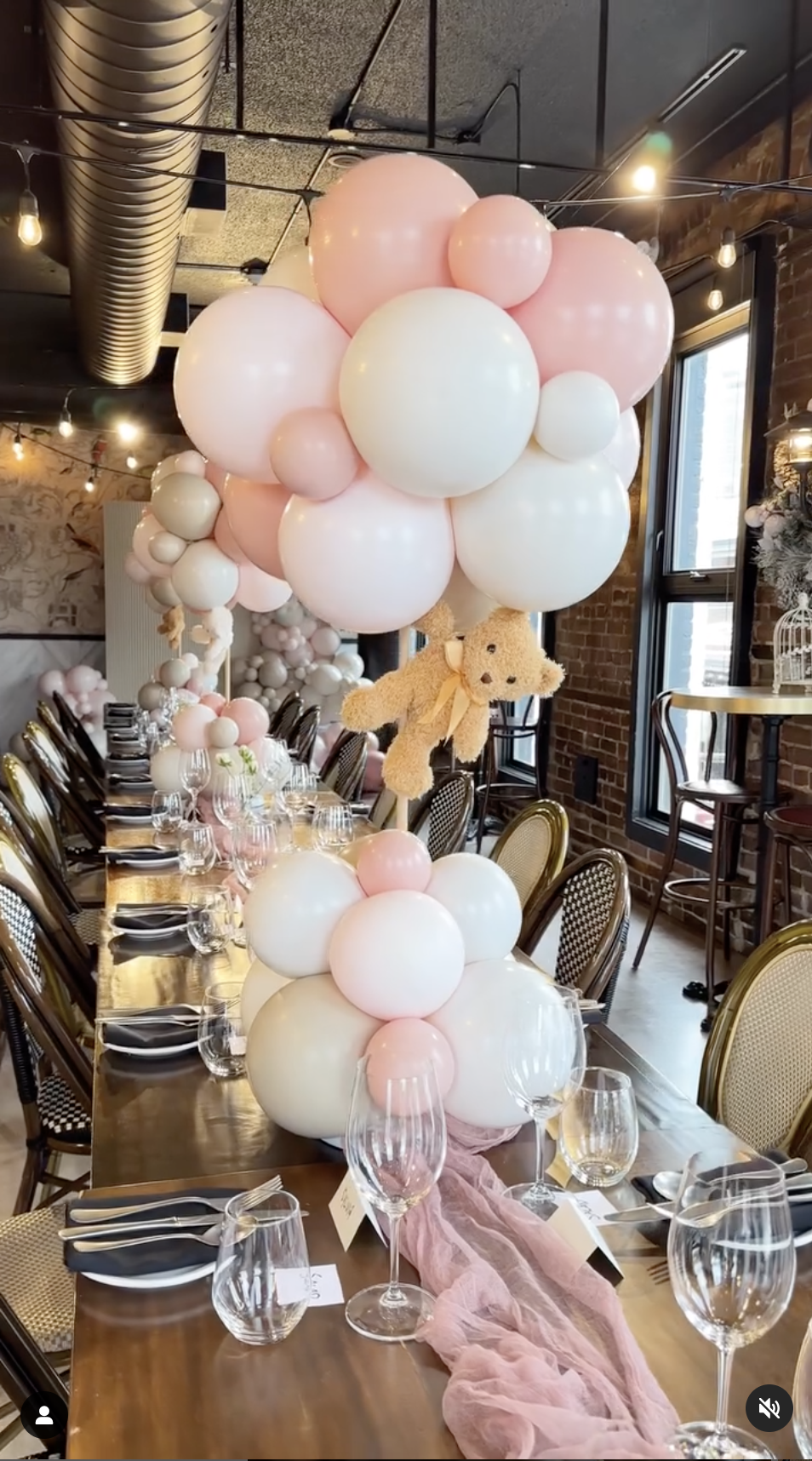 Indoor celebration with a long table decorated with pink and white balloons, teddy bear, and pink fabric runner, set for a meal with glassware, plates, and cutlery.