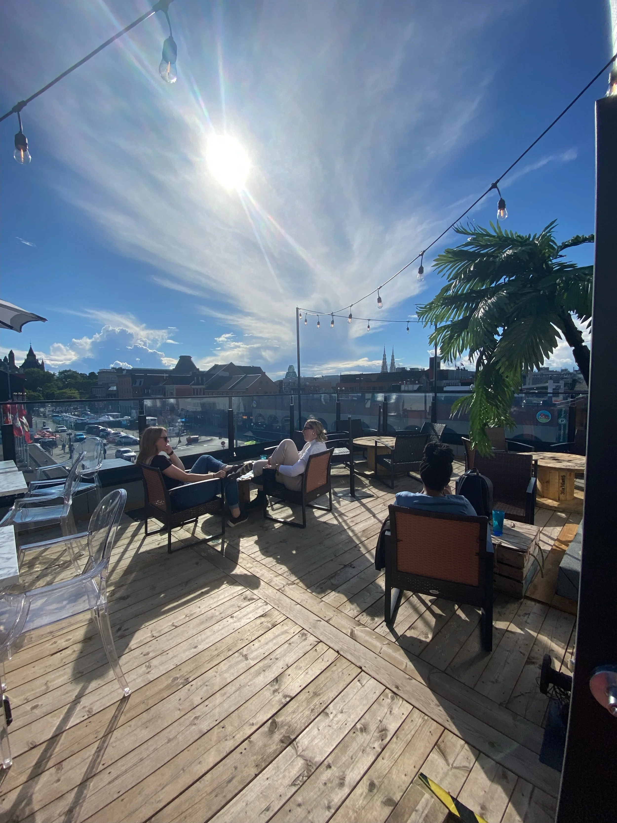People sitting and chatting on an outdoor terrace with string lights, a potted plant, and views of a city skyline under a bright, partly cloudy sky. rooftop, Ottawa, venues, byWard Market Ottawa events
