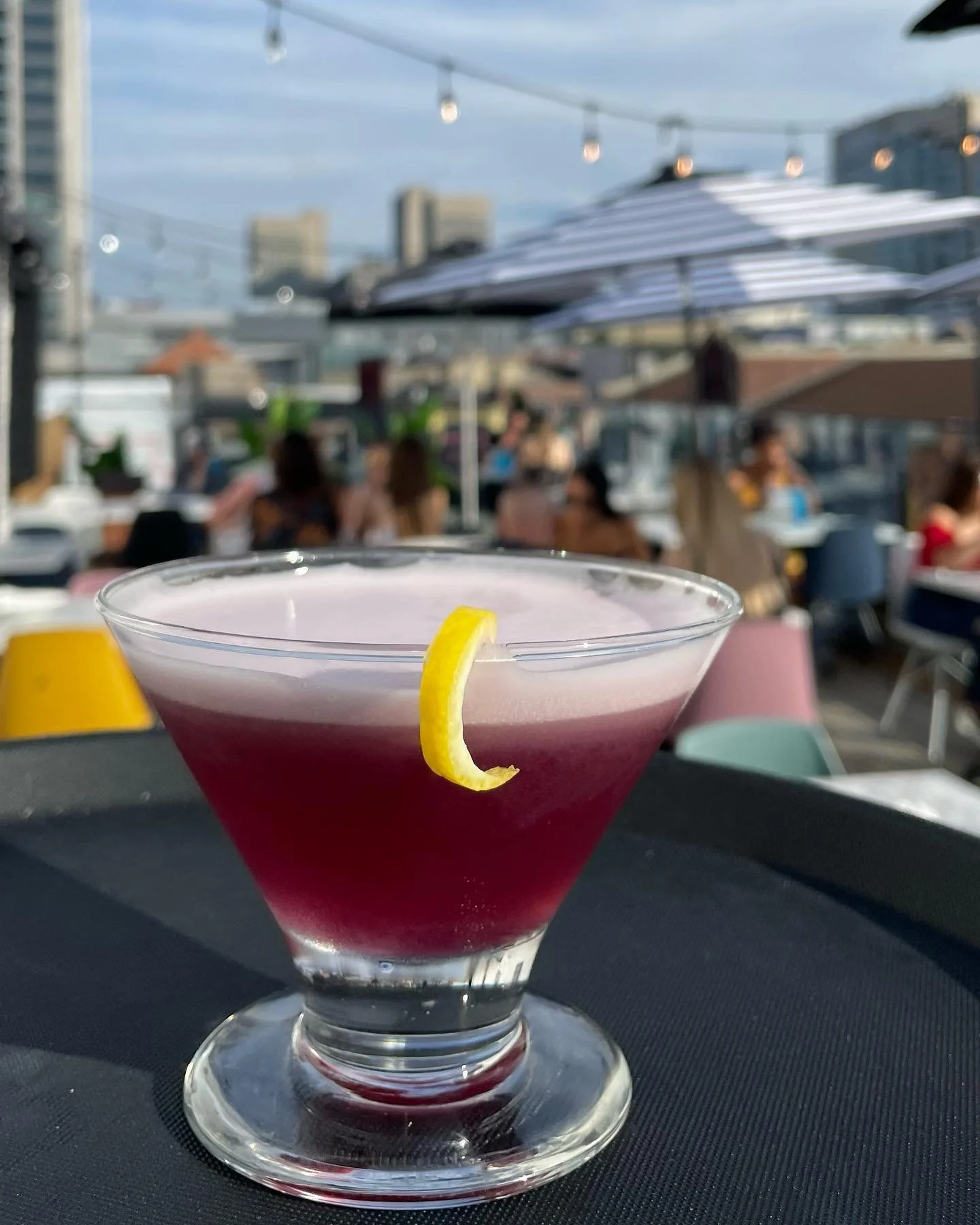 A pink cocktail with a lemon twist garnish on a black table outdoors at a rooftop bar, with people and city buildings in the background.