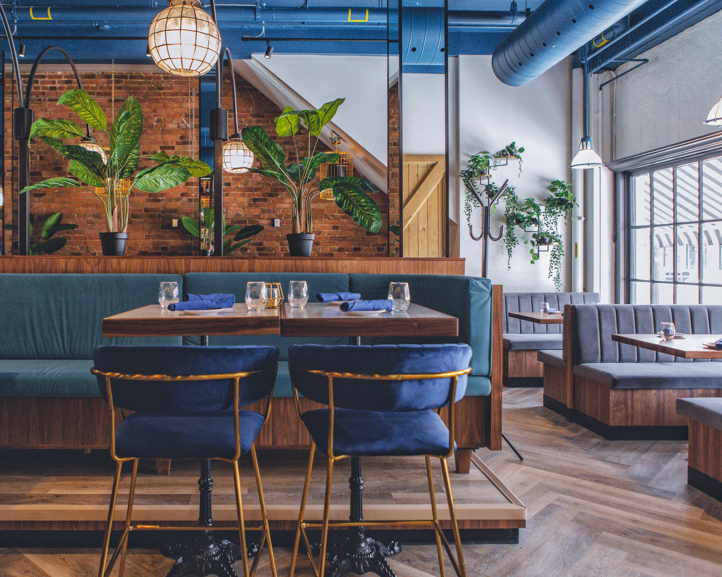 Interior of a modern restaurant featuring exposed brick walls, green indoor plants, blue upholstered seating, and wooden tables set with glasses and napkins. Artistic lighting and large windows let in natural light.