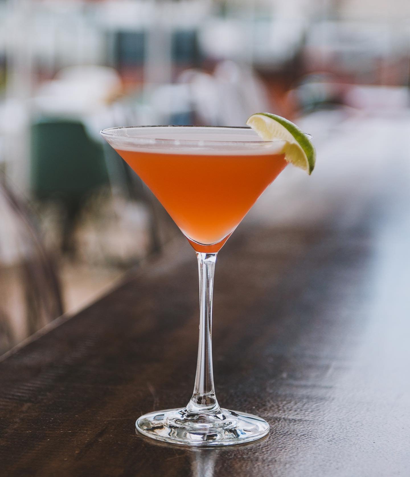 A pink cocktail in a martini glass garnished with a lime wedge, placed on a dark wooden surface. varied cocktails on our rooftop and a full menu. 