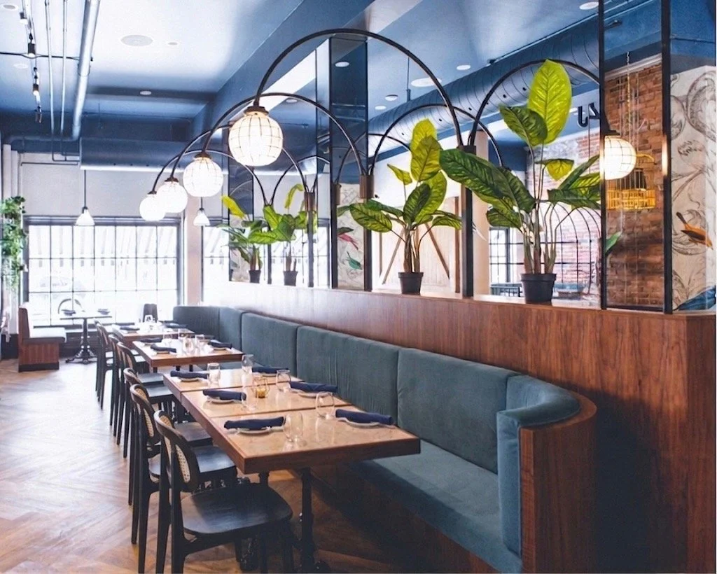 A modern restaurant interior with wooden tables set with plates, glasses, and napkins, a long teal upholstered banquette, decorative plants, and glass lanterns hanging from the ceiling. Bright natural light fills the space through large windows.