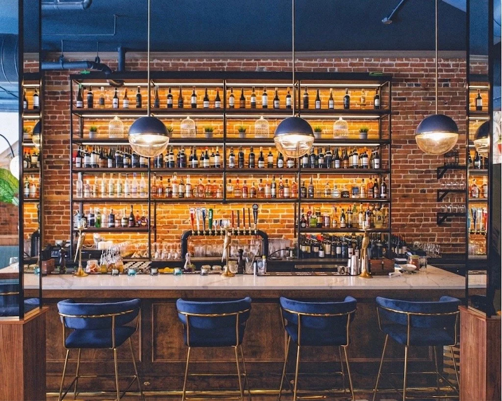 Modern bar with shelves full of bottles, illuminated with warm lighting, a brick wall background, black pendant lights, and four navy blue barstools at a white countertop. exposed brick wall, heritage building from 1875. a unique events venue