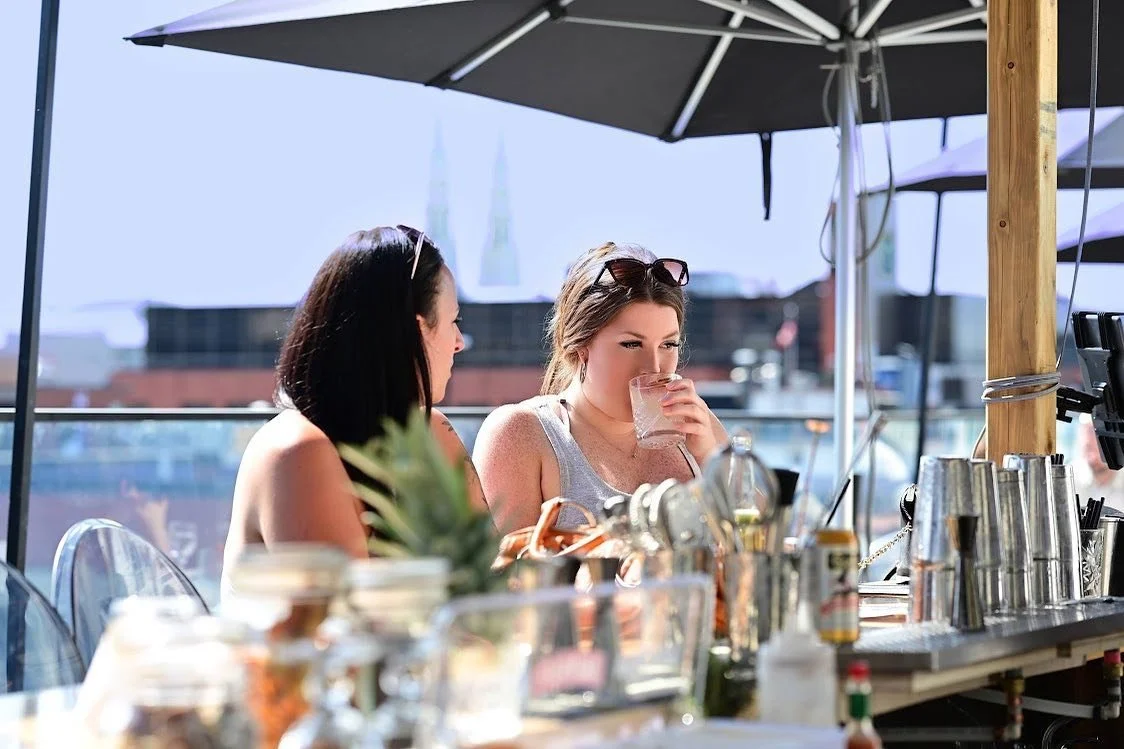 Two women sitting at an outdoor bar under umbrellas, one drinking from a glass, with a city skyline and clear sky in the background.