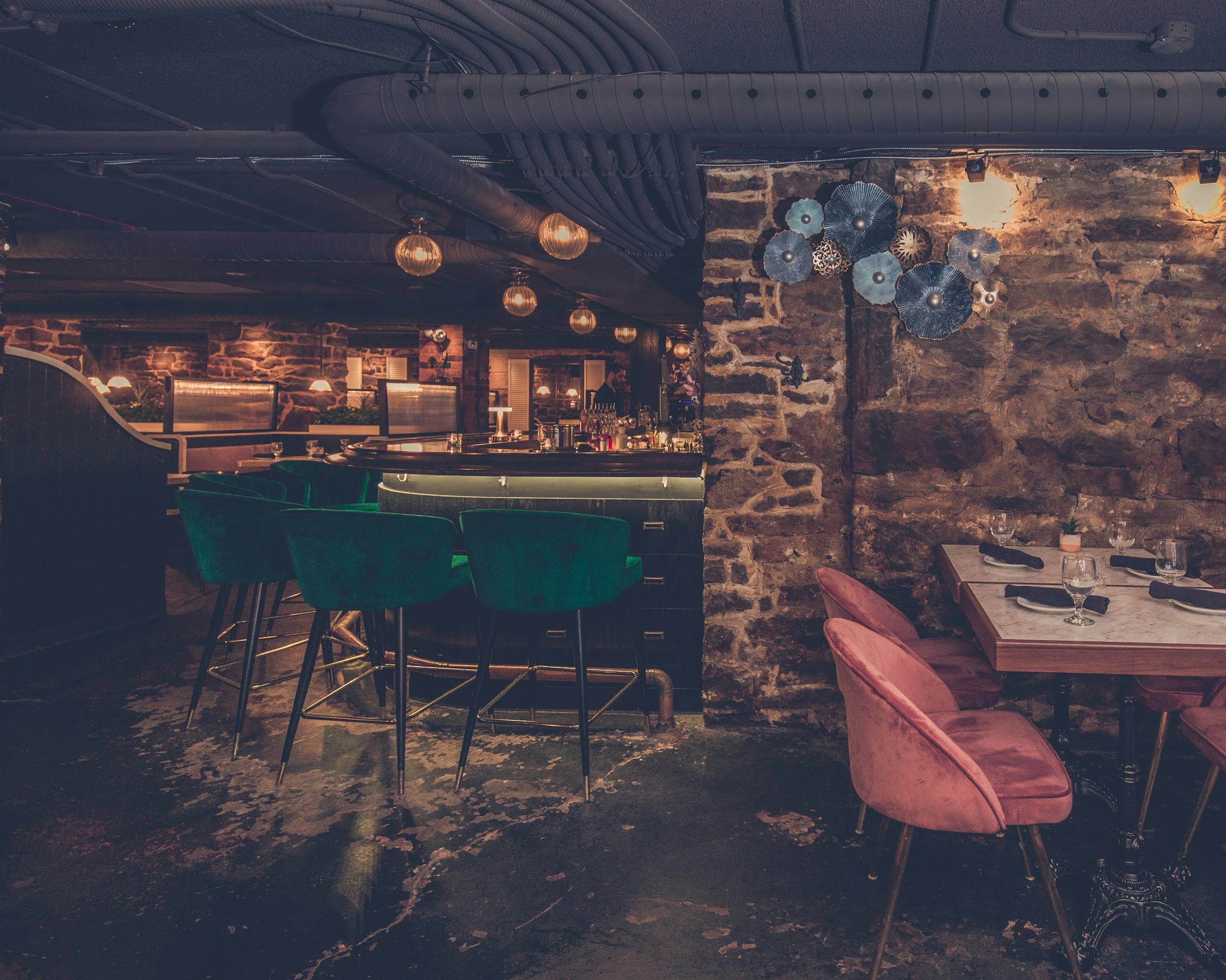 Interior of a restaurant or bar with exposed brick walls, modern hanging lights, and seating with teal and pink chairs. There is a decorative wall art installation of metallic flowers on one of the walls. Apothecary, heritage, stone walls, byWard