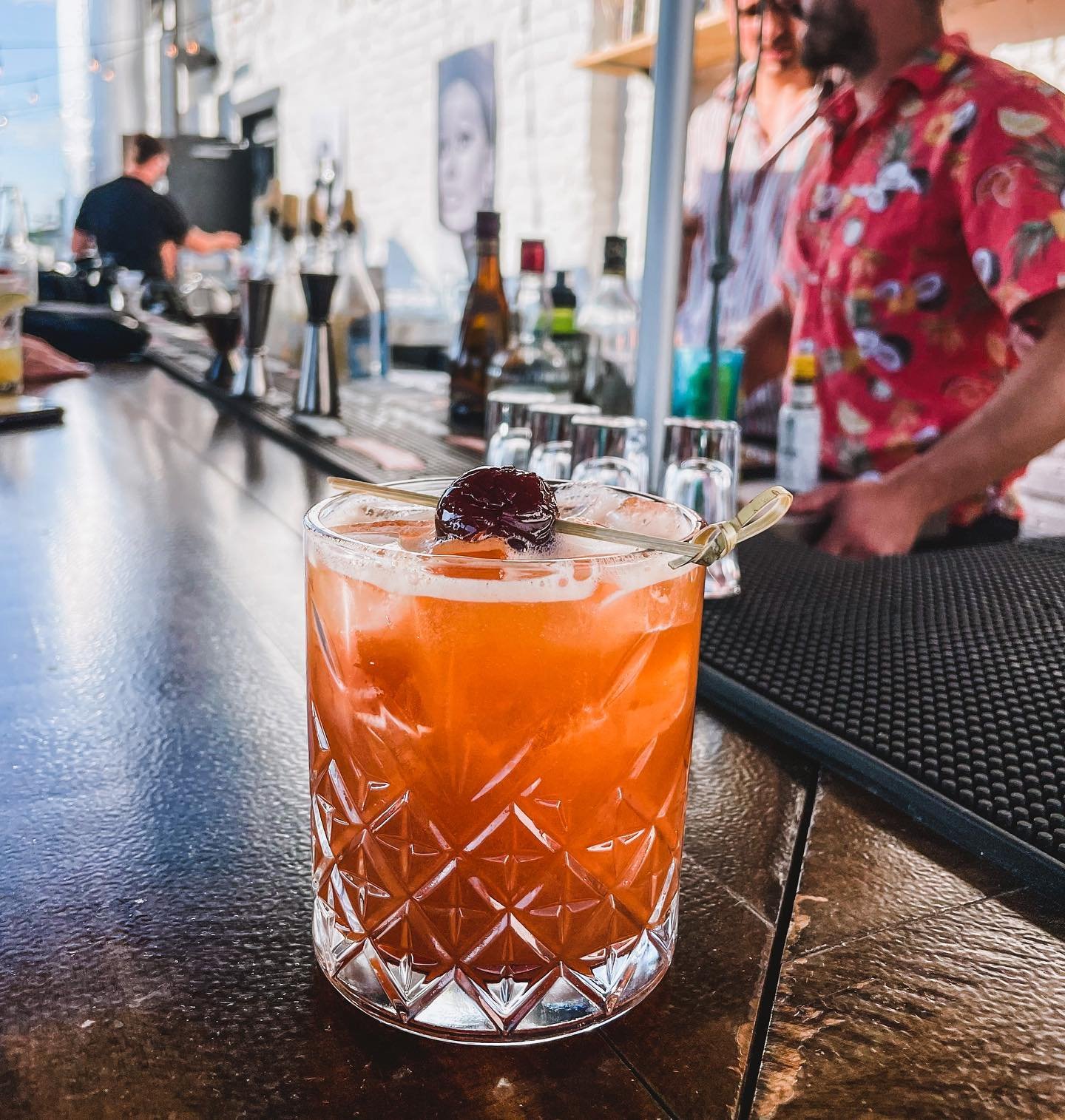 An orange cocktail with a cherry garnish on a bar counter with bartenders and bottles in the background.