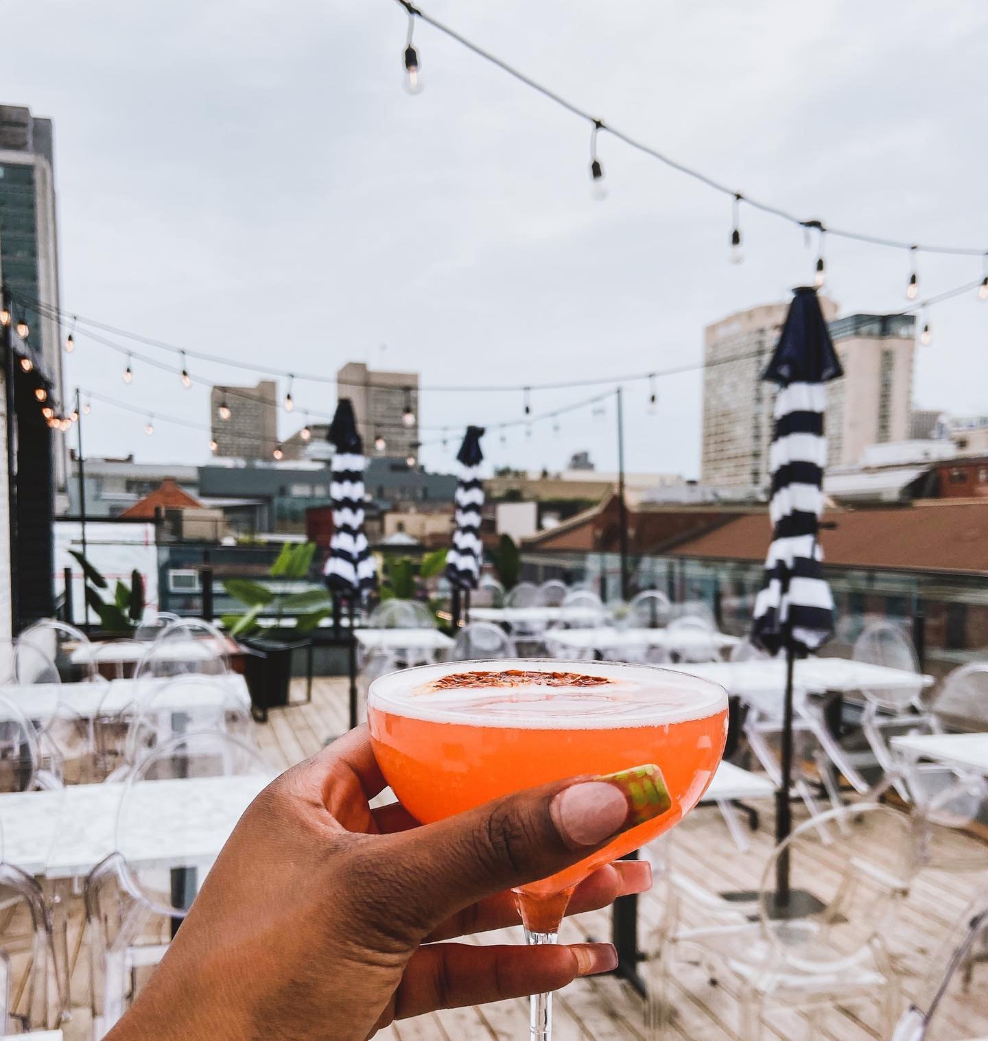 A person holding a cocktail in an outdoor rooftop setting with string lights, umbrellas, chairs, and tables, city buildings in the background during daytime.
