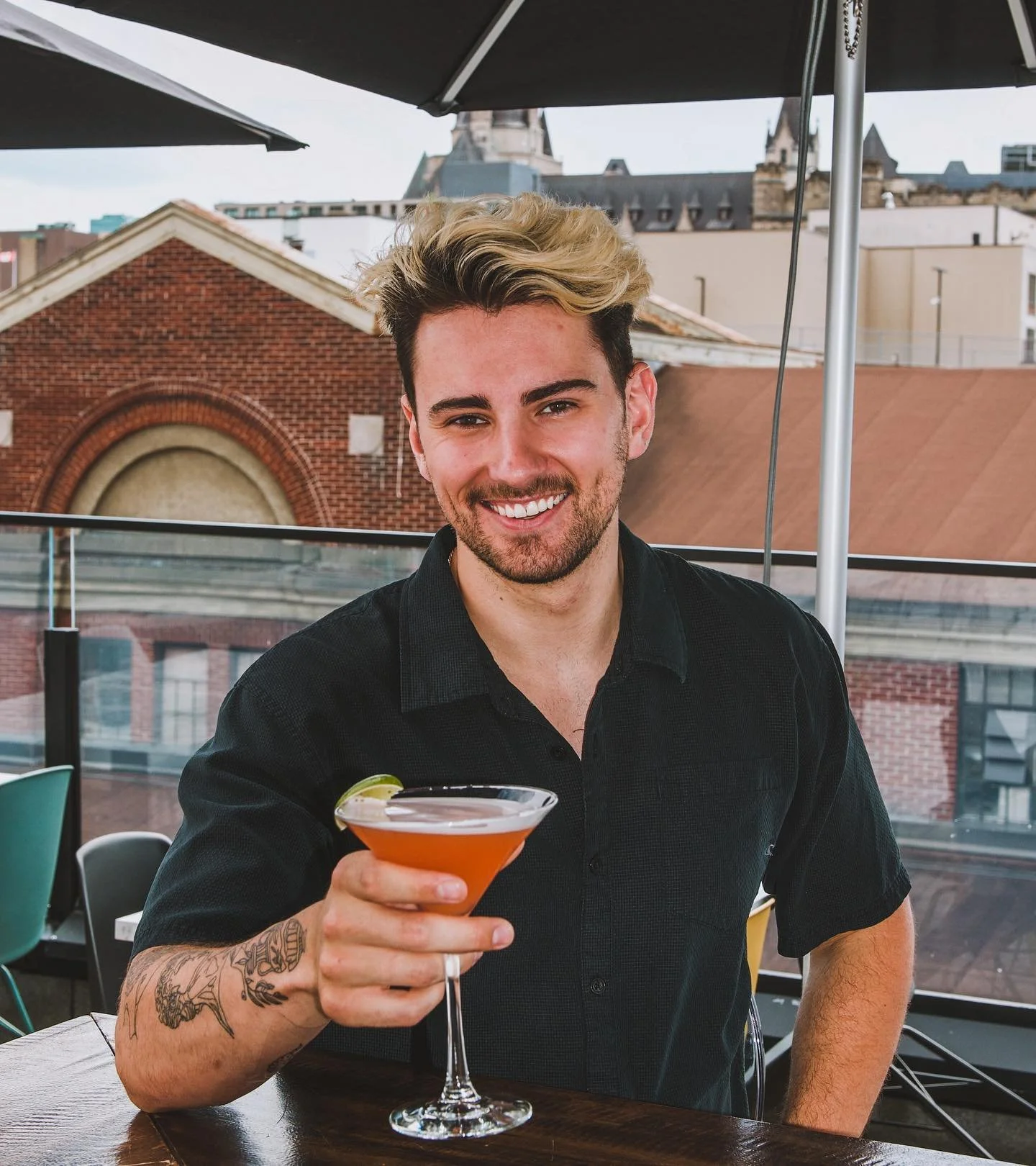 A smiling man with styled hair and a beard holding a cocktail glass with an orange drink and lime wedge at outdoor restaurant or bar. rooftop vibes. byWard Market Ottawa best venues private events, birthday, new spots, coolers spots in Ottawa