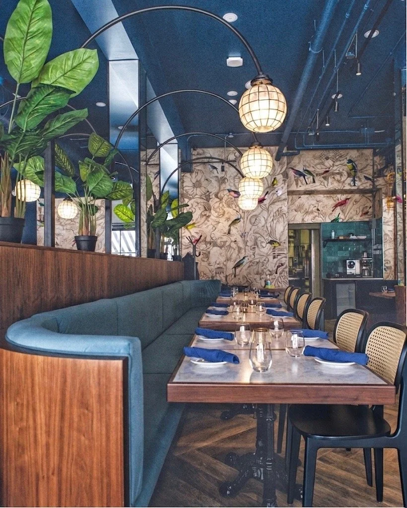 A modern restaurant interior with a long wooden table set with blue napkins and glasses, a teal cushioned bench, chairs with cane backs, decorative plants, and hanging spherical pendant lights.