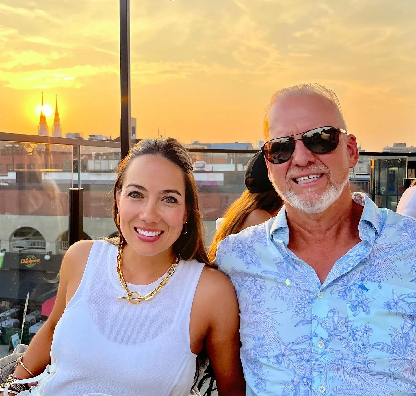 A woman and a man smiling at sunset during a rooftop gathering with a cityscape and church steeples in the background. Byward Market Views and vibes, best restaurant and rooftop. copper sights. 