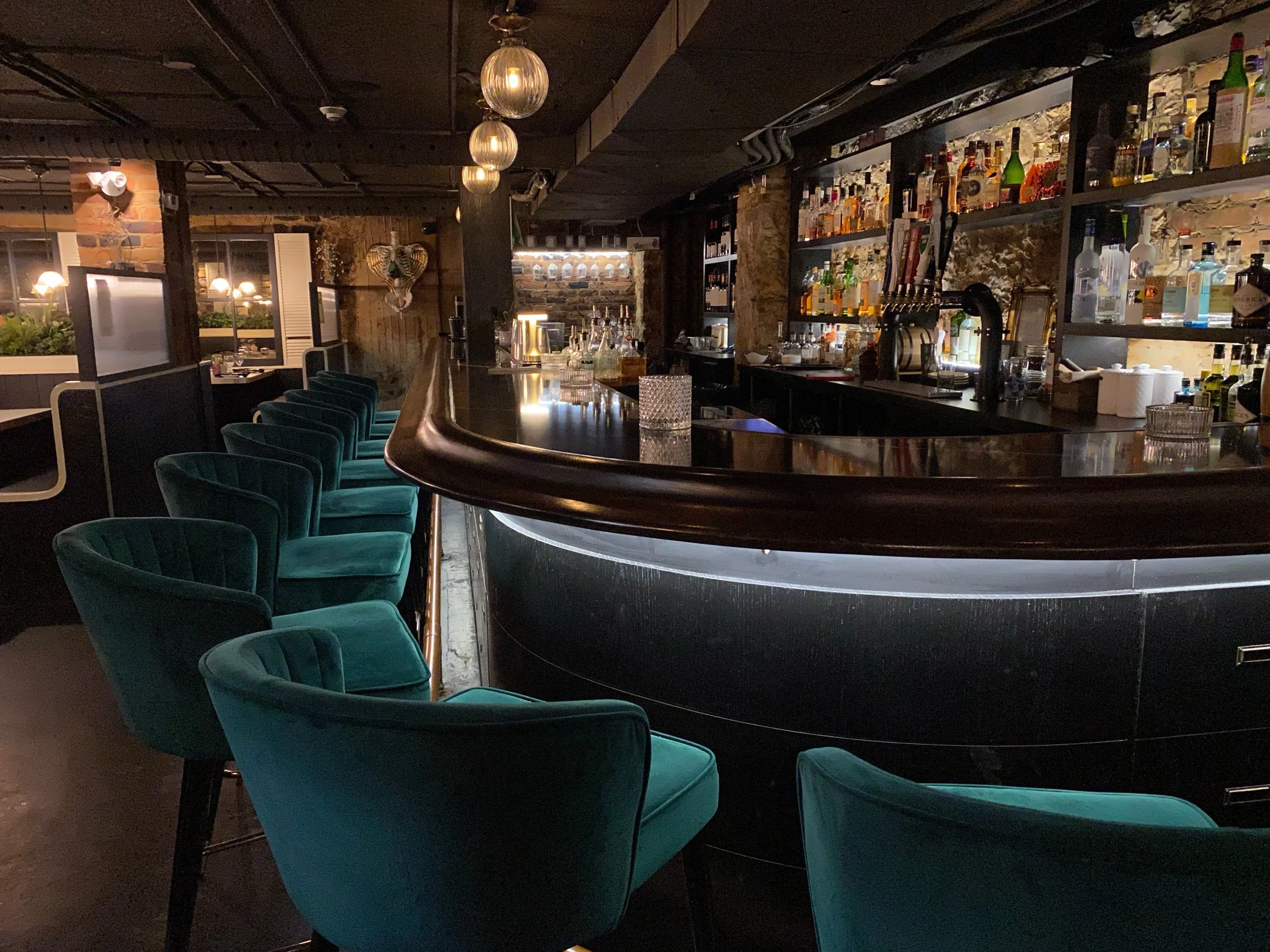 Inside a dimly lit bar with a curved dark counter and blue velvet chairs. Shelves behind the bar display various bottles of alcohol. Pendant lights hang from the ceiling, and a heart-shaped decoration is visible on the brick wall in the background.