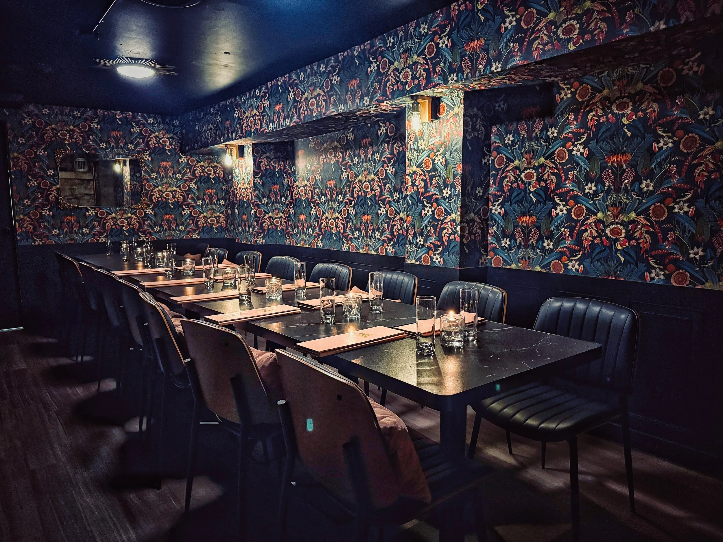 A dimly lit restaurant interior with long tables set for dining, glasses, and place settings. The walls are covered with dark, vibrant floral wallpaper, and there are wall-mounted lights illuminating the space.