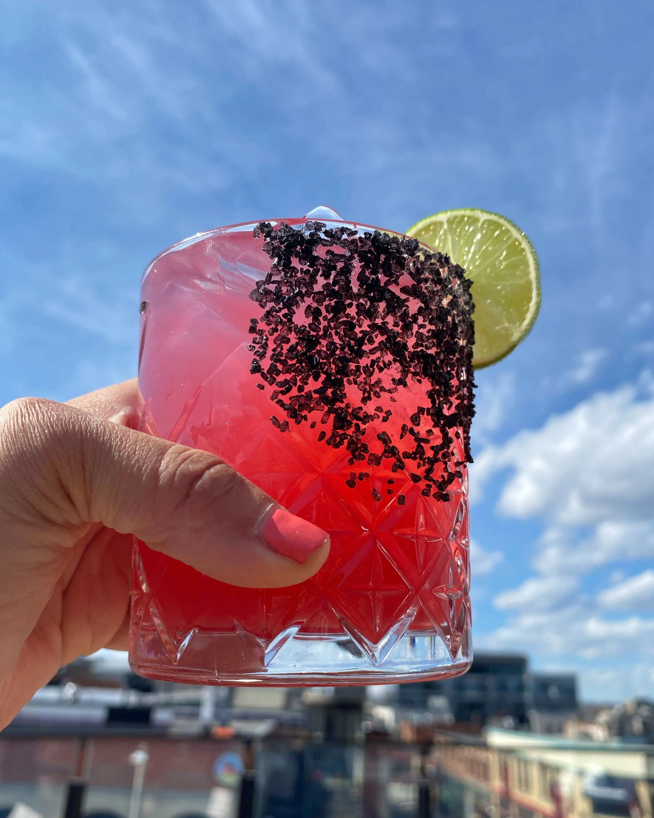 A pink-colored drink in a clear plastic cup with a black sugar rim and a lime wedge on the rim, held up against a blue sky with some clouds, likely outdoors.