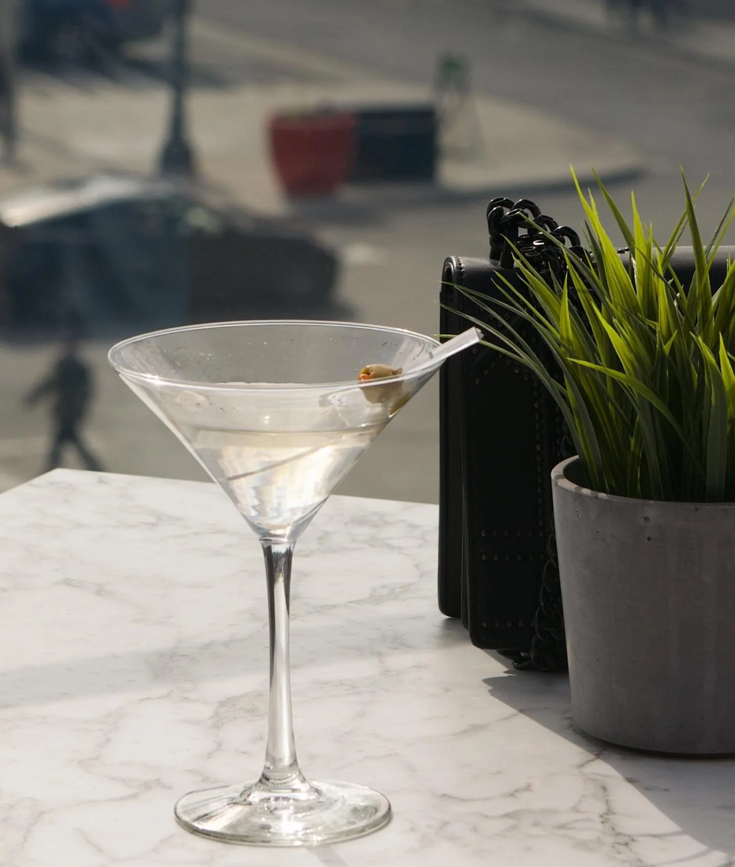 Empty martini glass with a small olive on a marble table, with a potted plant and a black object in the background.