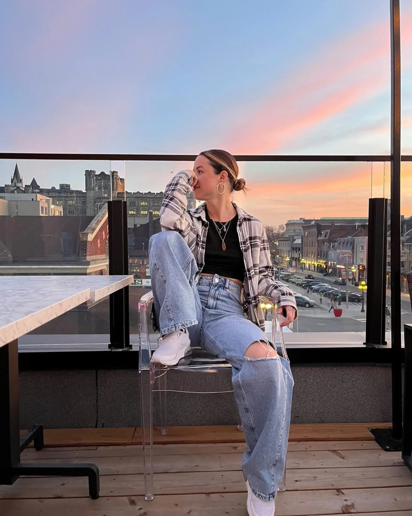 A woman with short hair tied in a bun, wearing large hoop earrings, layered necklaces, a black top, a plaid shirt, and ripped jeans, sitting on a transparent chair on a rooftop balcony during sunset, with a cityscape in the background.