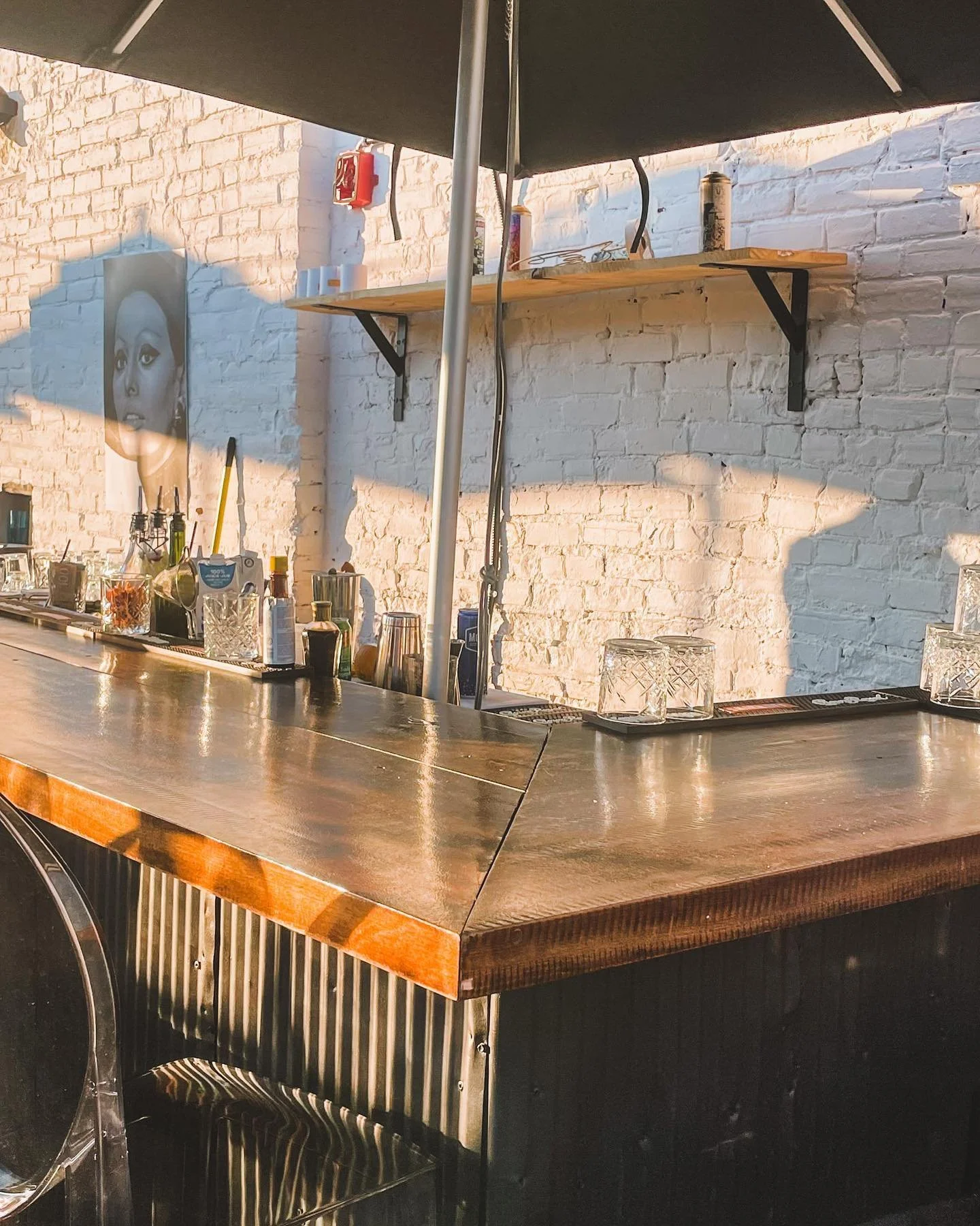 Part of a bar counter with glasses, bottles, and bar tools, against a white brick wall with shelf and artwork, under a large black umbrella. built in bar rooftop, Ottawa, byward market copper sights, sunsets and views