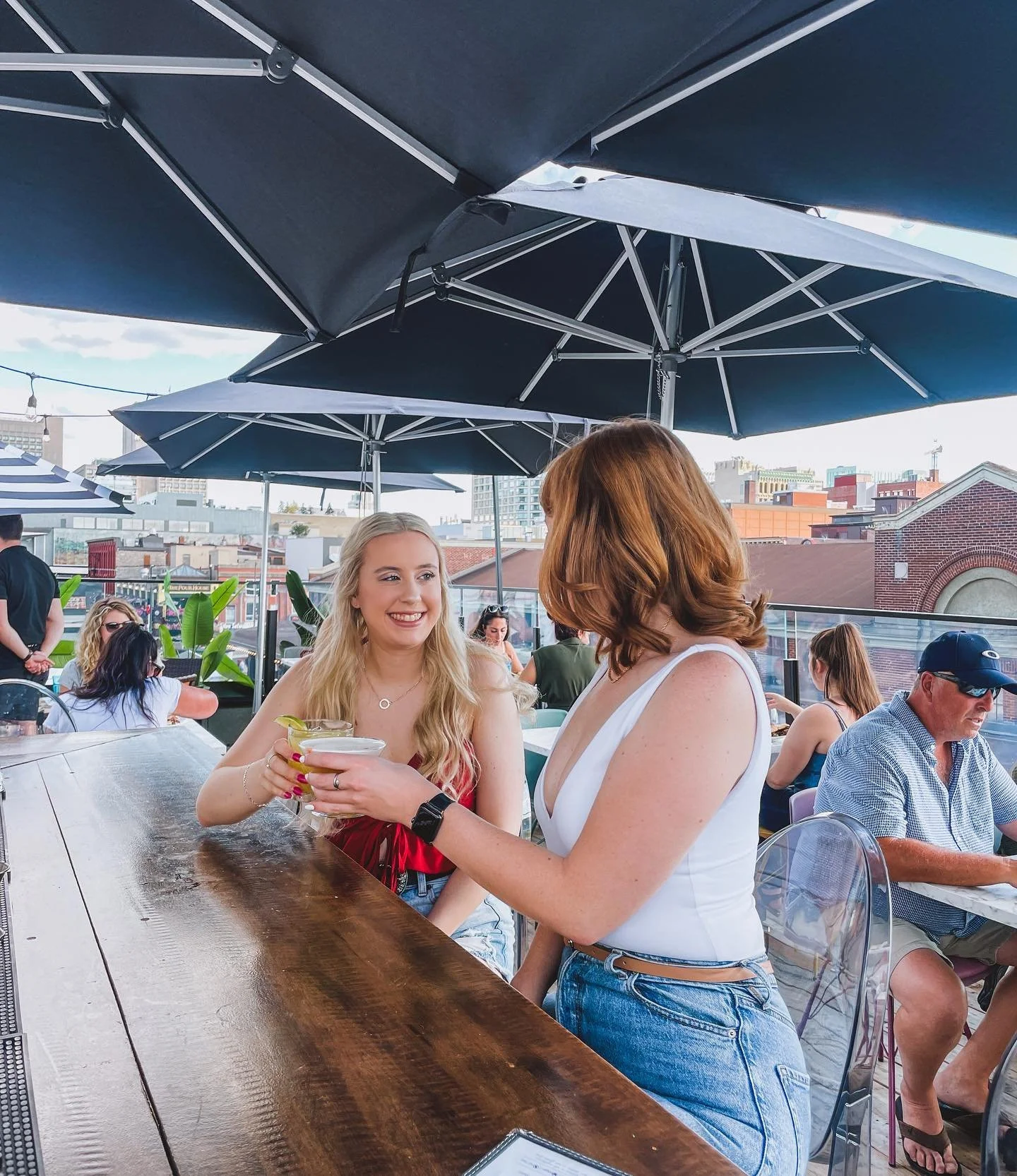 Two women are socializing under large black umbrellas at an outdoor rooftop restaurant or bar. One woman with blonde hair in a red top is holding a cocktail and smiling at the other woman with reddish-brown hair in a white sleeveless top. Other peopl