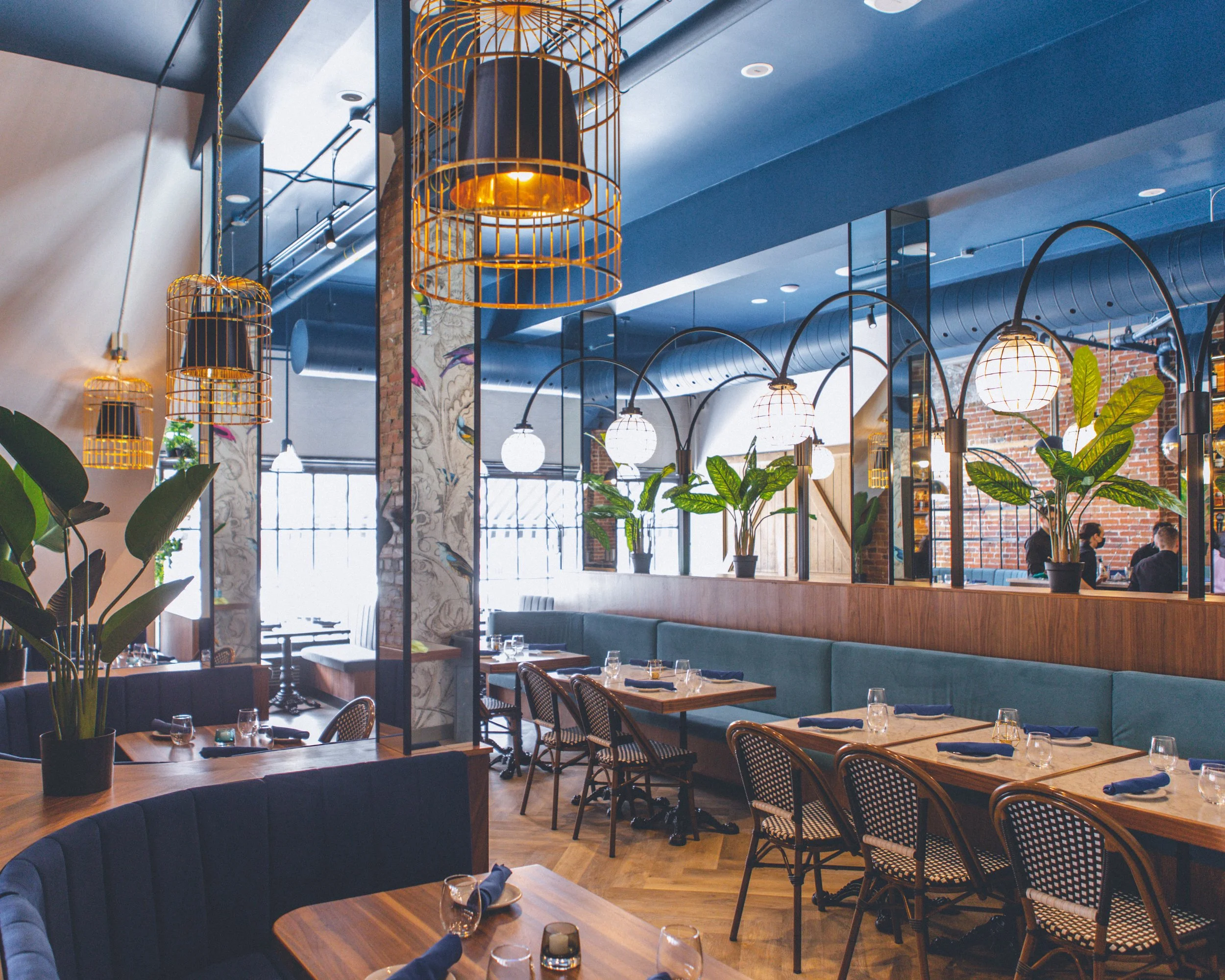 Interior of a restaurant with wooden tables, large windows, greenery, exposed brick walls, artistic wall decor, and hanging bird cage lamps, creating a modern and cozy ambiance. the velvet booths are warm and inviting. the space is bright