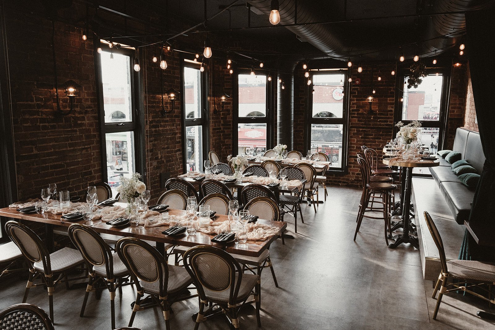 Interior of a cozy restaurant or event space with exposed brick walls, large windows, string lights, and tables decorated with flowers and tableware for a gathering or celebration.