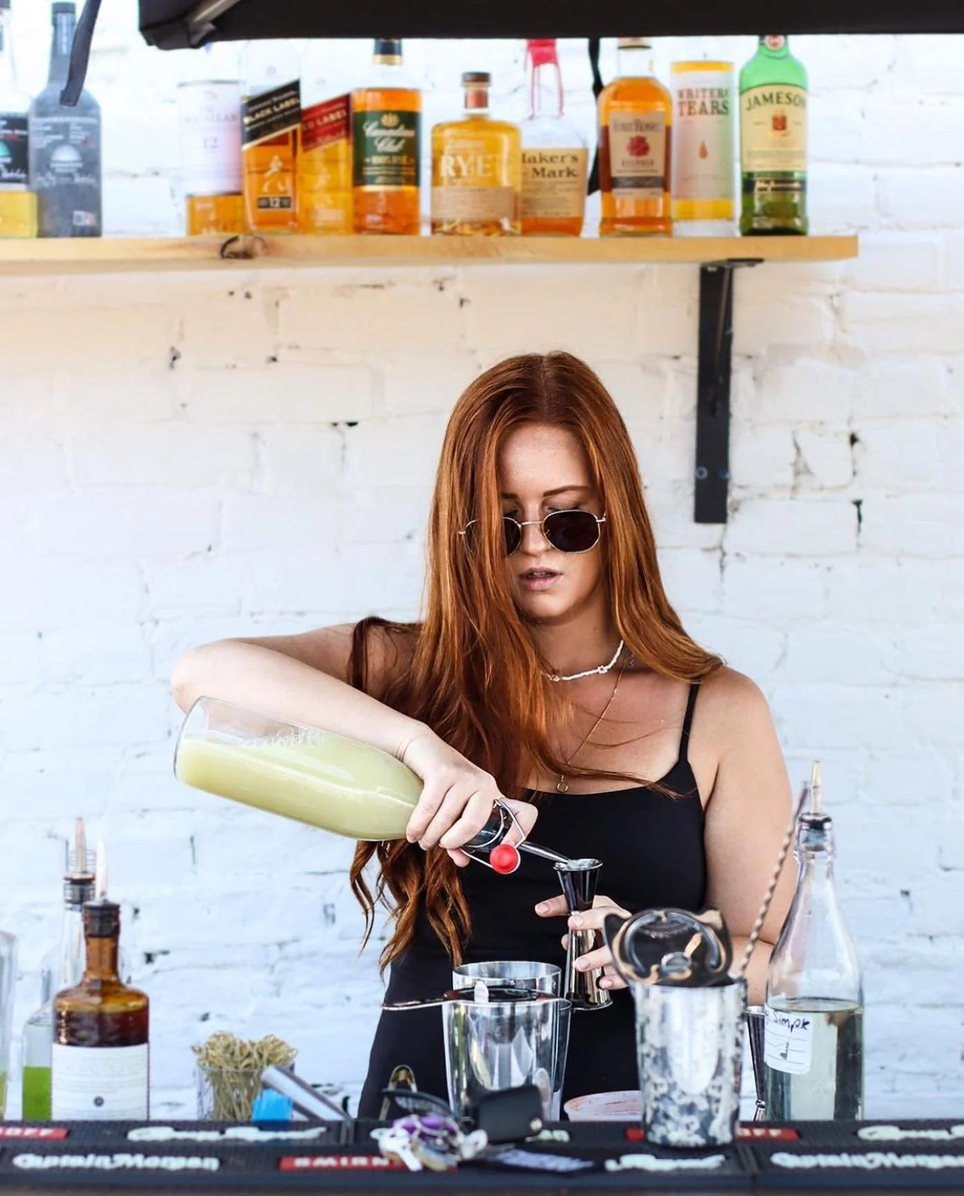 A woman with long red hair, wearing sunglasses and a black top, pouring a green cocktail from a blender into a glass in a bar setting with various bottles and bar tools on the table and shelves in the background.