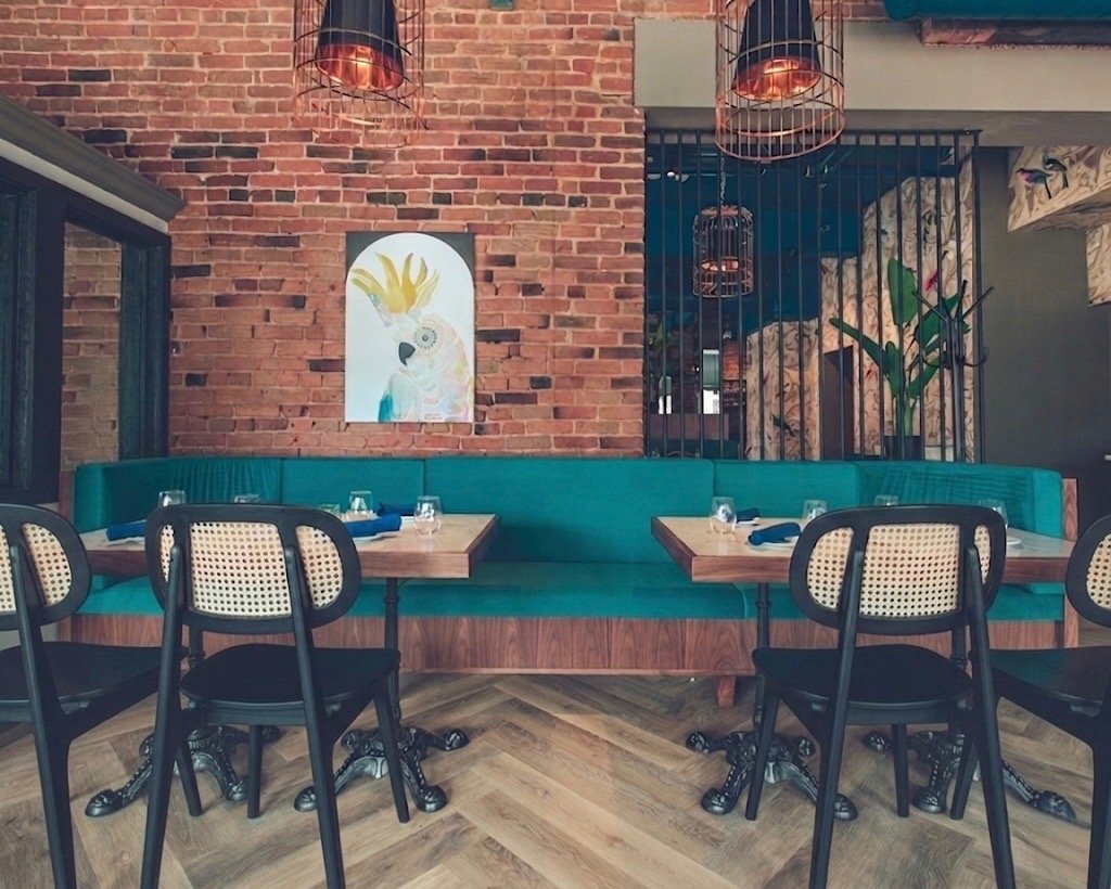 Interior of a restaurant with turquoise velvet booth seating, wooden tables, brick exposed wall with bird artwork, hanging birdcage light fixtures, and a decorative plant. a perfect backdrop for an intimate gathering private event