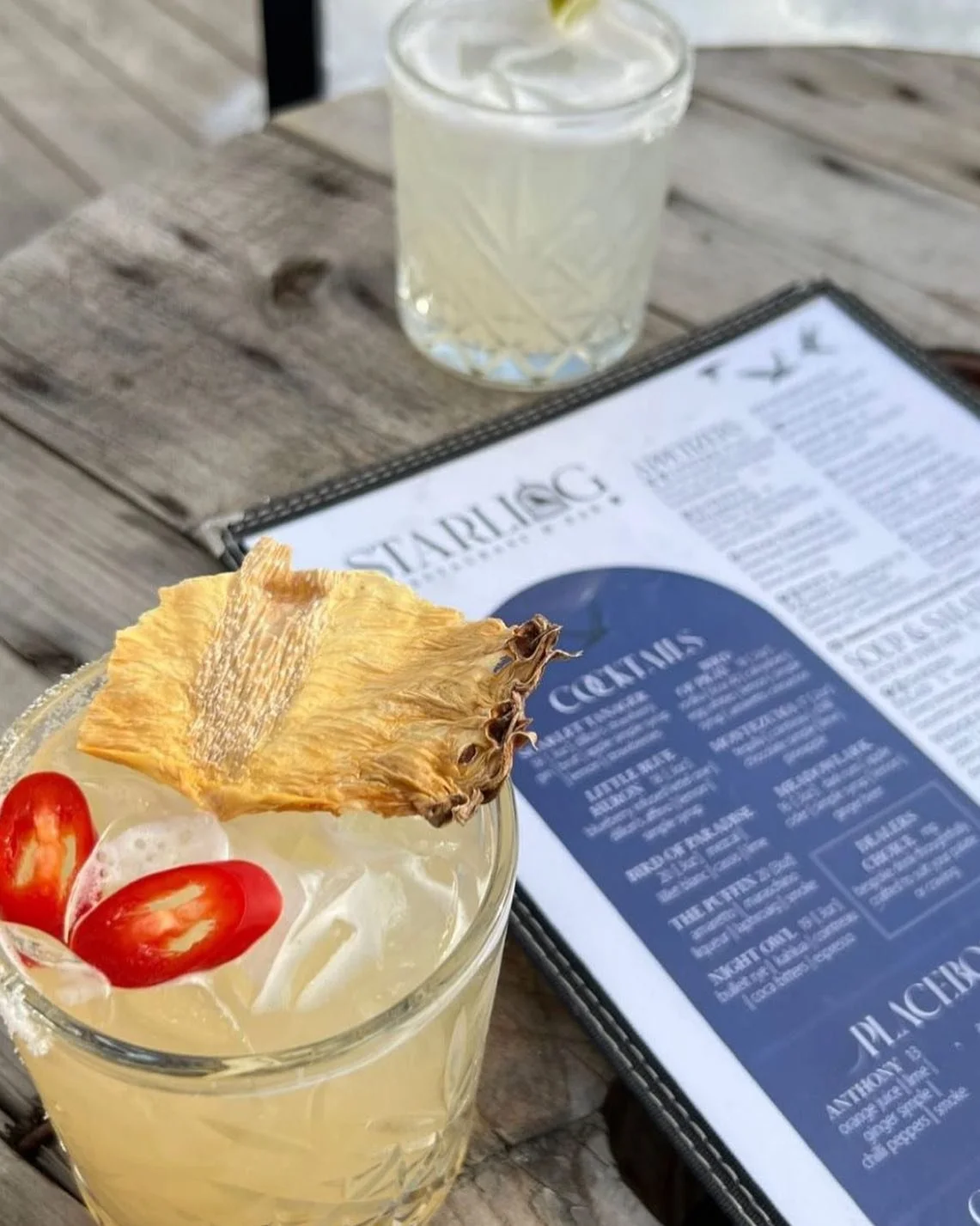 A cocktail in a glass garnished with dried pineapple slices and cherry tomatoes, with a menu and another drink in the background on a wooden table.