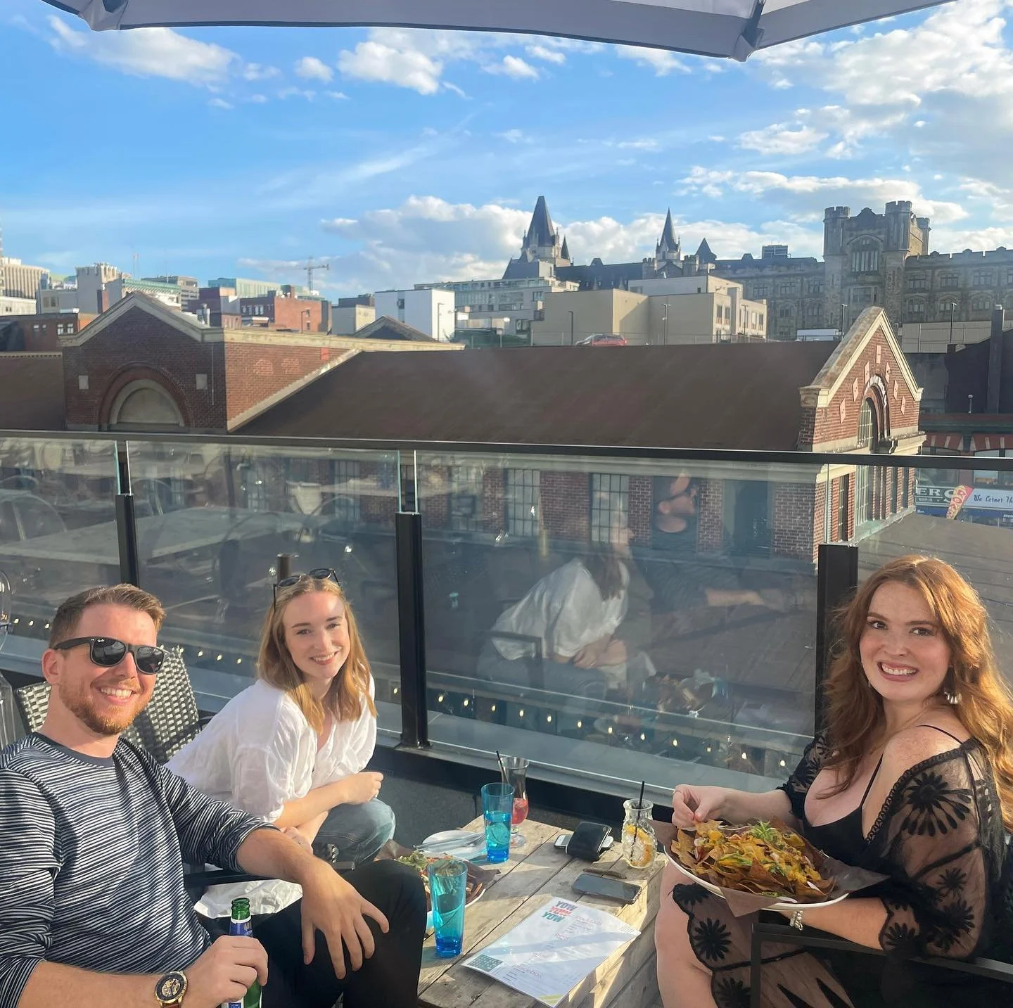 Four people sitting at an outdoor rooftop restaurant with a city skyline in the background, enjoying food and drinks on a sunny day.