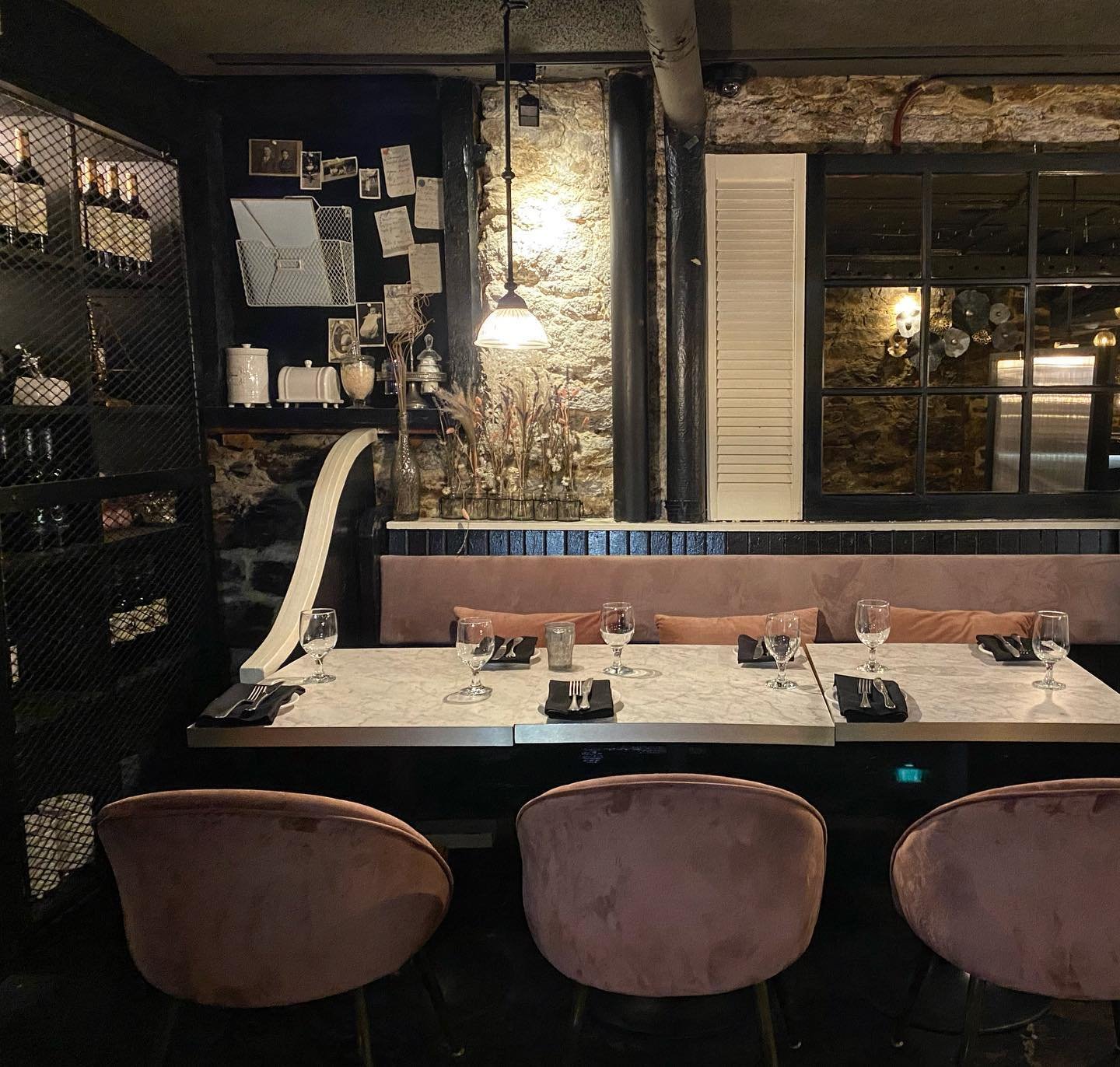 A cozy restaurant dining area with a long marble table set for five, with black napkins, silverware, and wine glasses. Pastel pink chairs line the front; the backrest of the booth is pink. The wall features a combination of exposed brick and black pa