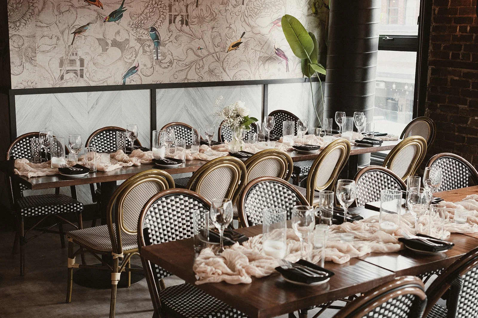 A restaurant dining area with a long table set for a meal. The table has a beige cloth runner, black napkins, and glasses for water and wine. Several chairs with different patterns and finishes surround the table. A vase with white flowers and green 