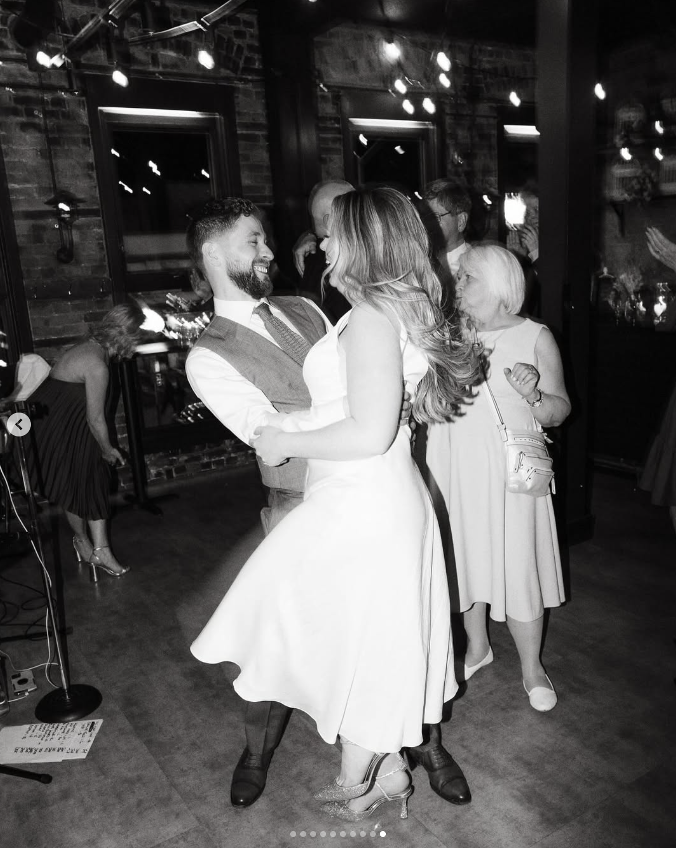 A black-and-white photo of a couple dancing closely at a social gathering or party, with joyful expressions. The woman is in a white dress with a full skirt, high heels, and has long hair. The man is wearing a vest, tie, and dress shirt. In the backg