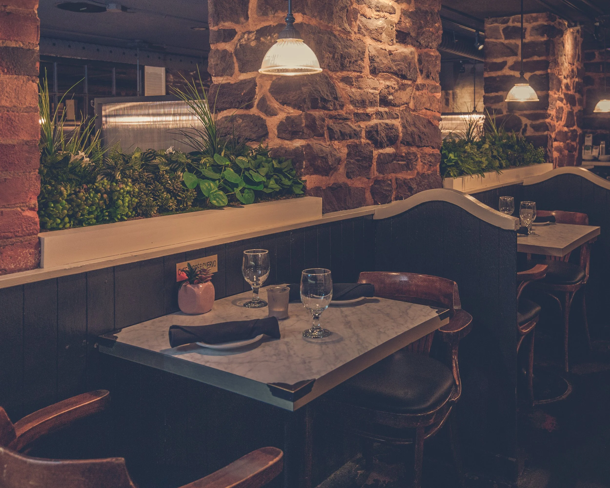 Inside a restaurant with brick pillars, hanging pendant lights, and a marble-top table set with wine glasses, napkins, and a small pink pot with plants. Apothecary Lounge, heritage building, timeless ambiance, modern vibes. decadent food. best bar