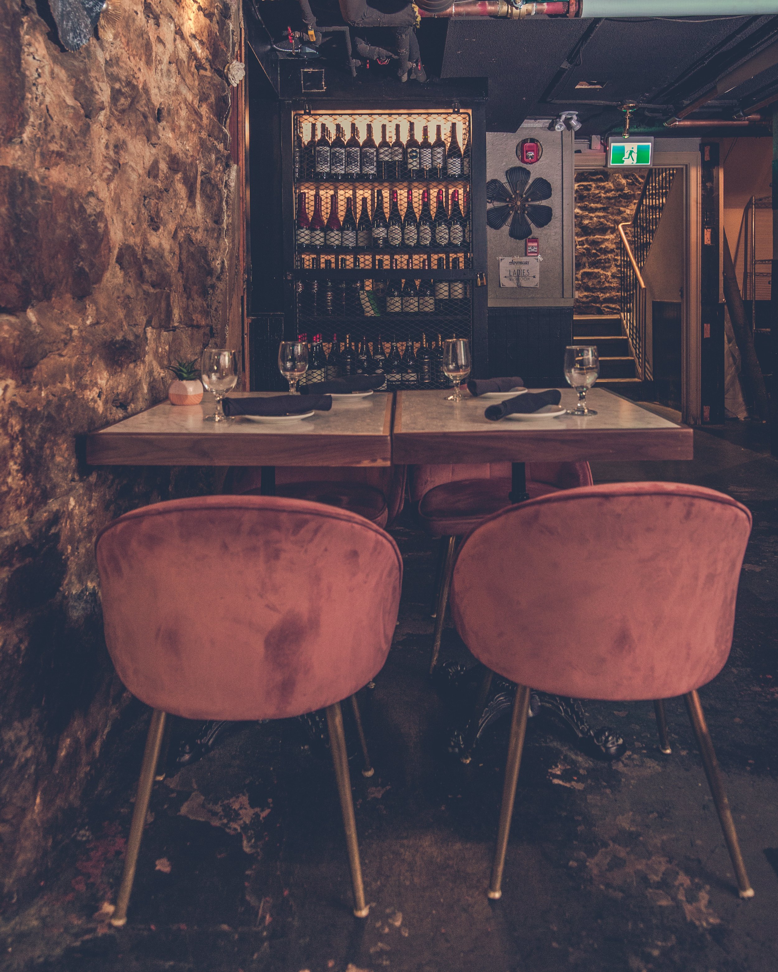 A restaurant table set with black napkins, empty wine glasses, and a small potted succulent plant in a pink pot. In the background, there is a wine bottle display encased in a black metal grid, exposed brick wall, and a staircase with an illuminated 