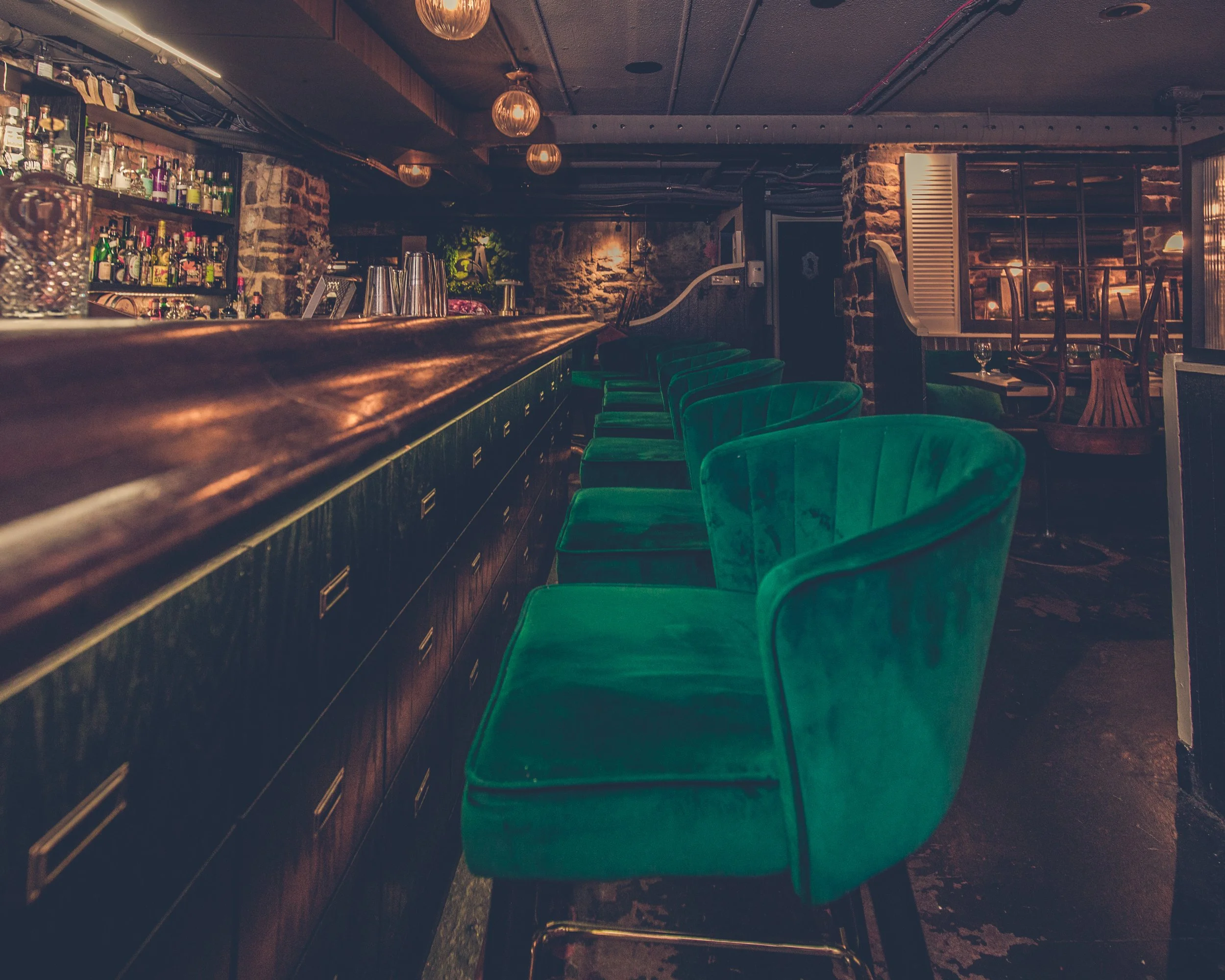 Interior of a dimly lit bar with a long wooden counter, green plush chairs, and shelves stocked with liquor bottles. Exposed brick walls and hanging light fixtures create a cozy ambiance.