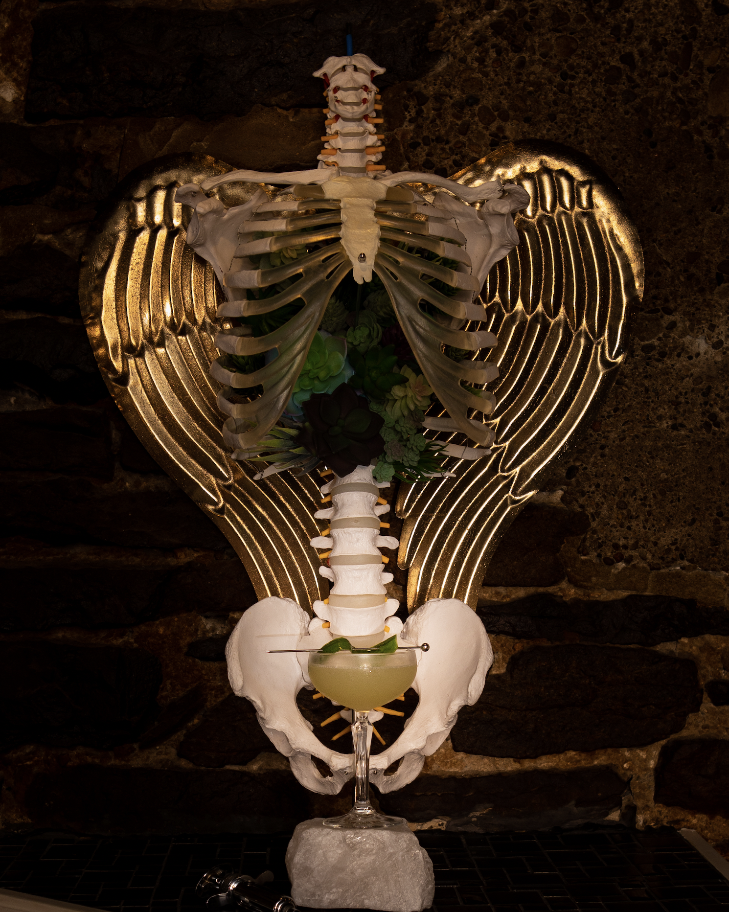 An artistic skeleton with angel wings holding a green cocktail in front of a dark textured background.