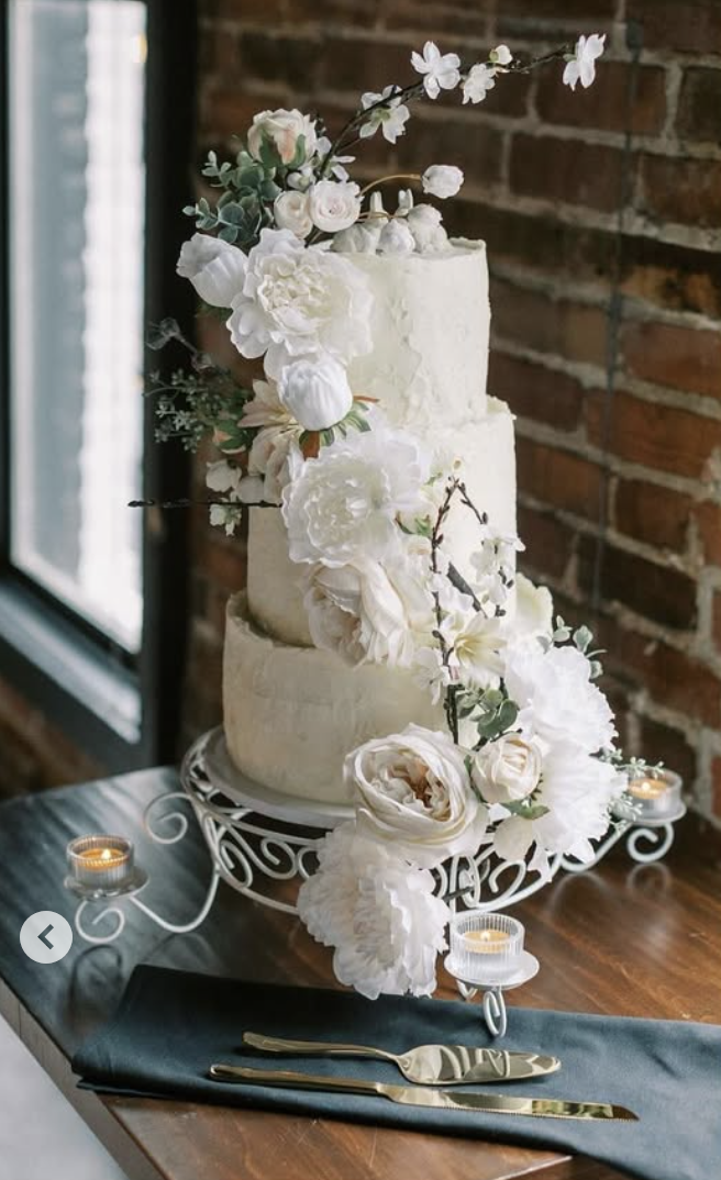 Three-tiered white wedding cake decorated with white flowers and greenery, placed on a decorative stand on a dark table with teal napkin and gold utensils, against a brick wall background.