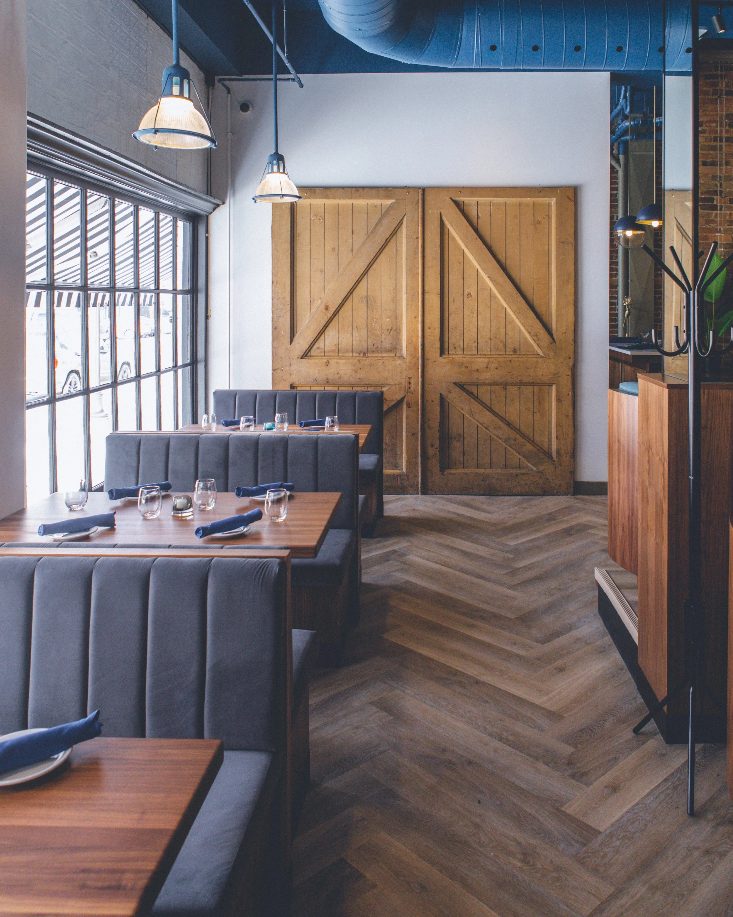 Interior view of a restaurant with large window, wooden sliding barn doors, wooden tables set with glasses and napkins, upholstered chairs, and industrial light fixtures with exposed ductwork and brick wall sections.