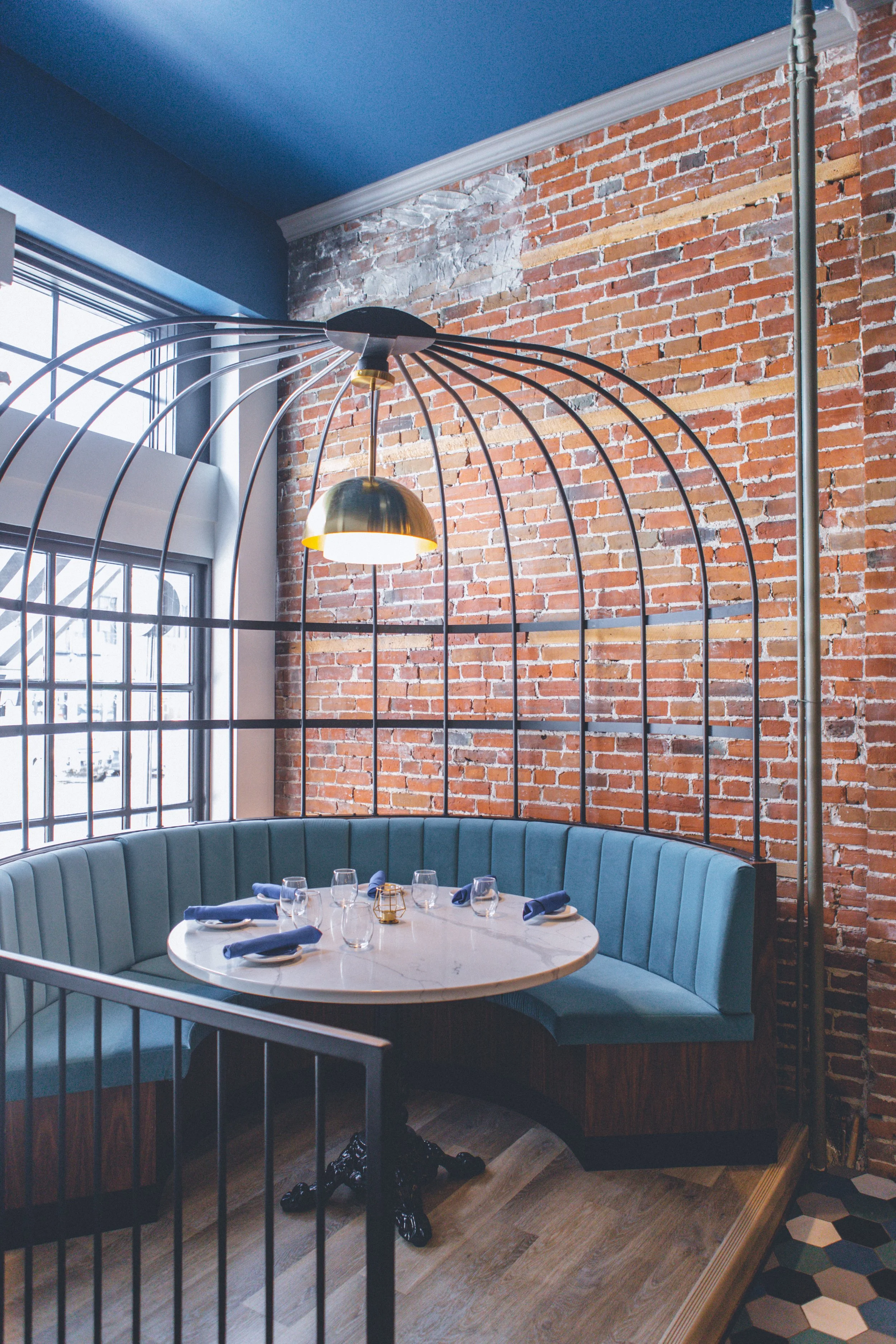 A round dining table with blue napkins, glasses, and a small golden candle holder, surrounded by a curved blue upholstered booth seat. The setting is inside a restaurant with exposed brick walls, wooden floor planks, a window with metal bars, and a m