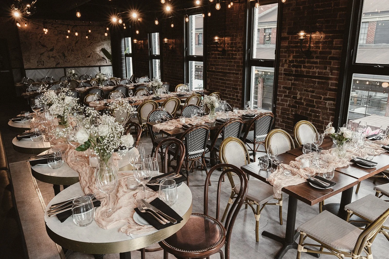 A restaurant or event space decorated for a special occasion with long wooden tables and smaller round tables, all set with elegant glassware, plates, black napkins, and pink cloth runners. Vases with white and blush flowers adorn the tables. The roo